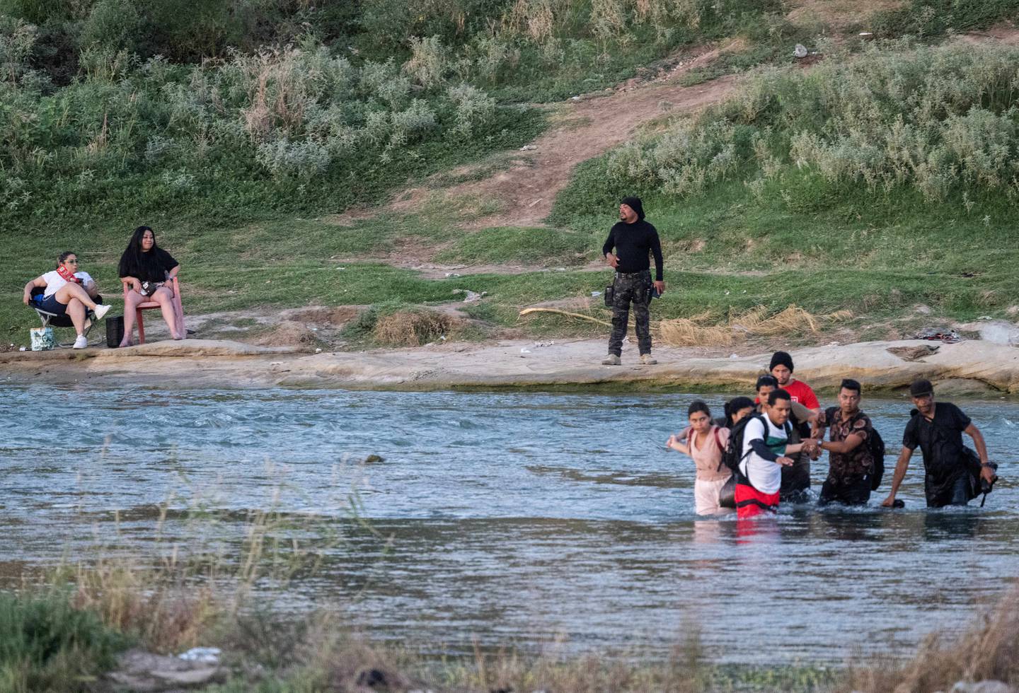 Dos mujeres (izq.) escuchando música y relajándose en la orilla del Río Grande observan a un grupo de migrantes que intentan cruzar el río hacia Eagle Pass, Texas,