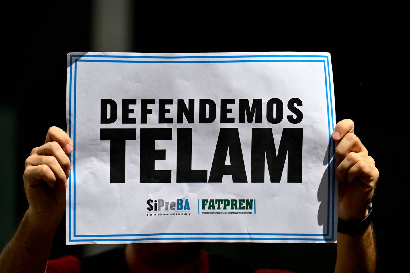 Un periodista sostiene un cartel que dice 'Defendemos Telam' afuera de la sede de la agencia de noticias estatal Telam durante una protesta mientras miembros de la Policía Federal Argentina custodian la entrada.