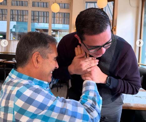 Imagen de Ignacio Santos con una camisa a cuadros azules sonriendo mientras saluda afectuosamente a Elías Alvarado quien viste una camisa de color negro.