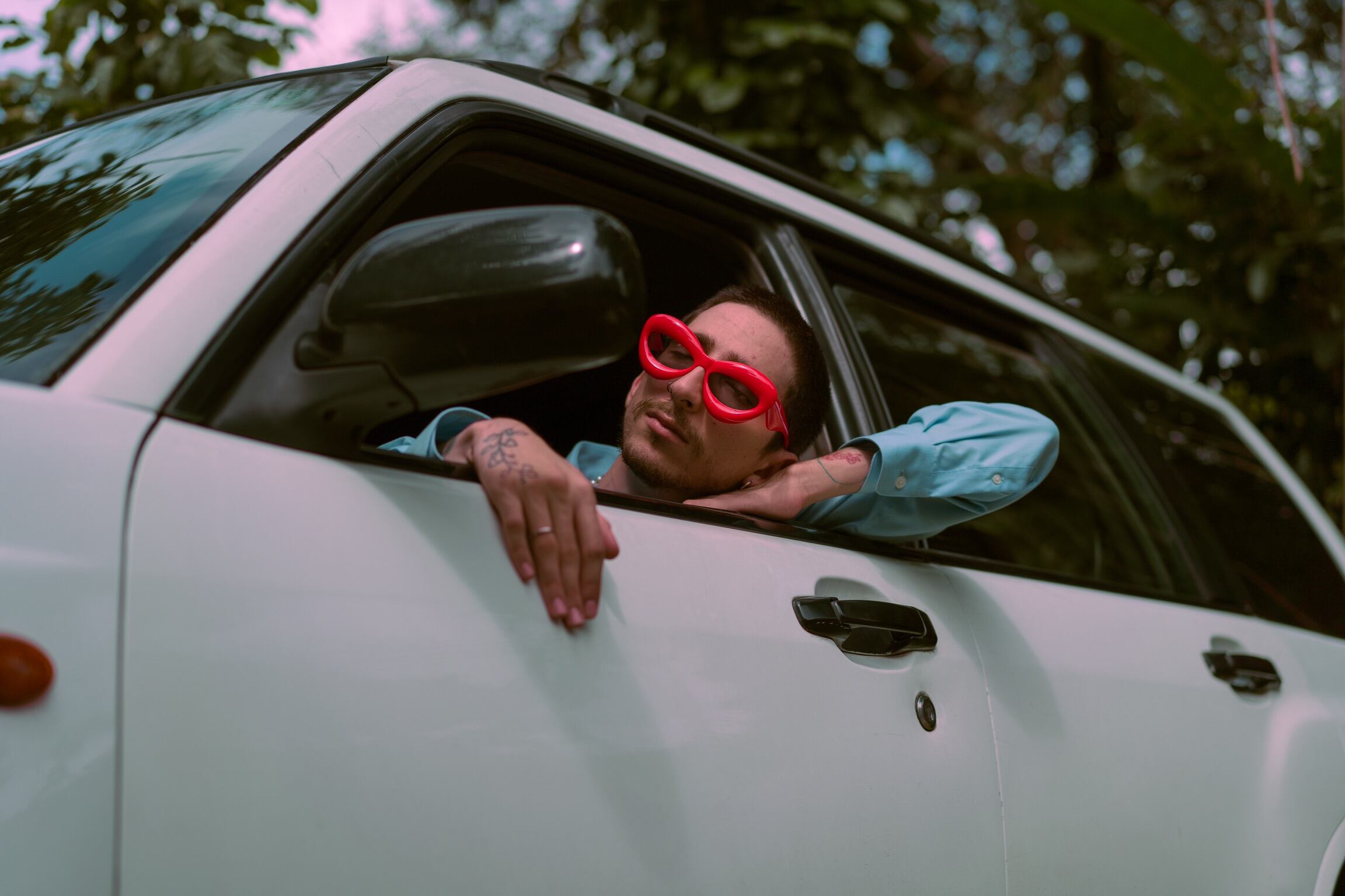 Un hombre dentro de un carro blanco. Se asoma por la ventana, usa lentes de color rojo.