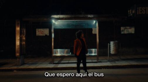 Hombre de pie en una parada de autobús iluminada en la noche, simbolizando reflexión y crítica sobre temas sociales.