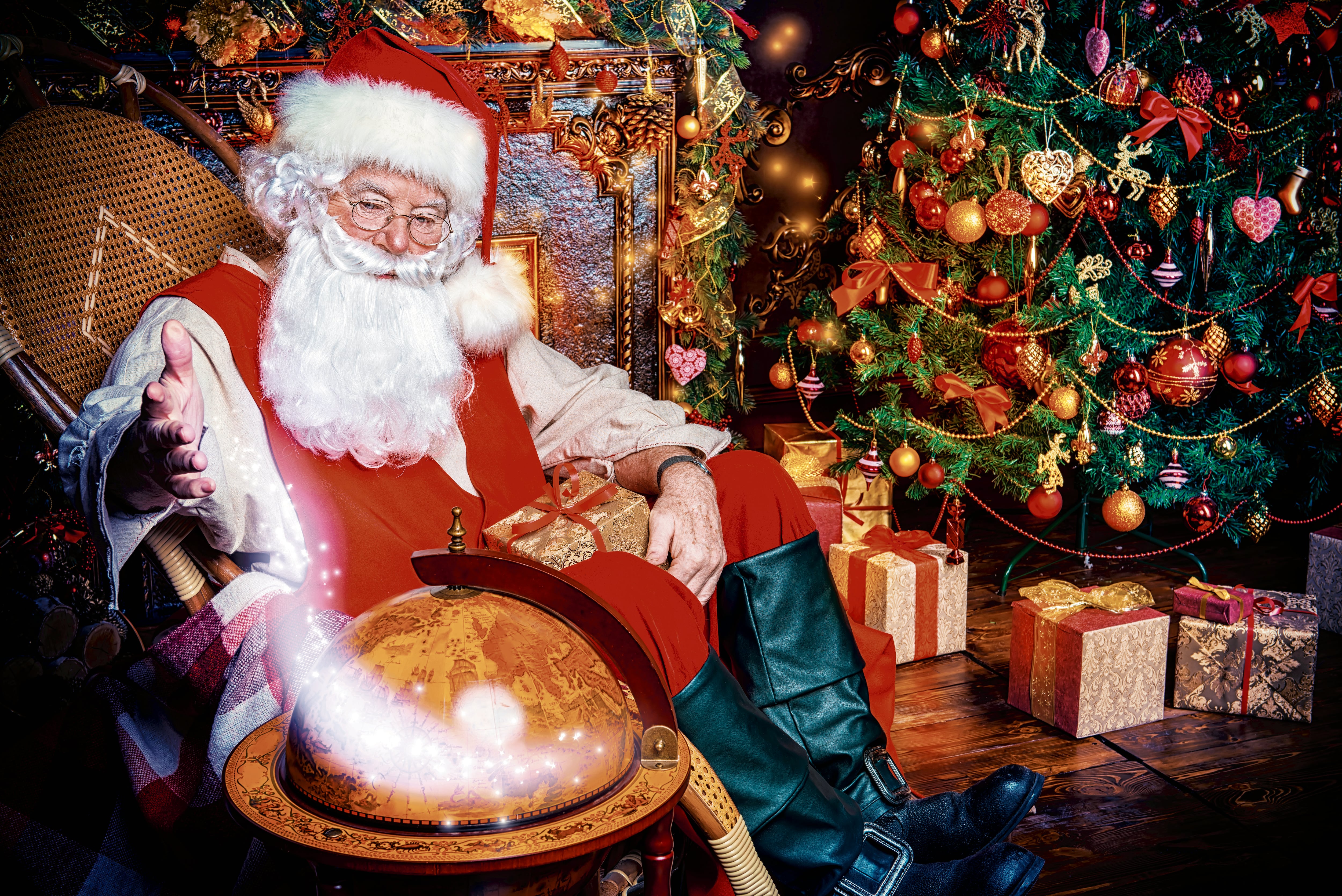 Santa Claus vestido de rojo y blanco señala un globo terráqueo, con un árbol de Navidad decorado al fondo.