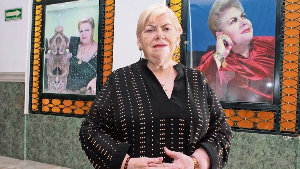 Imagen de la cantante Paquita la del Barrio vestida de negro, con su cabello rubio y posando frente a fotografías suyas.