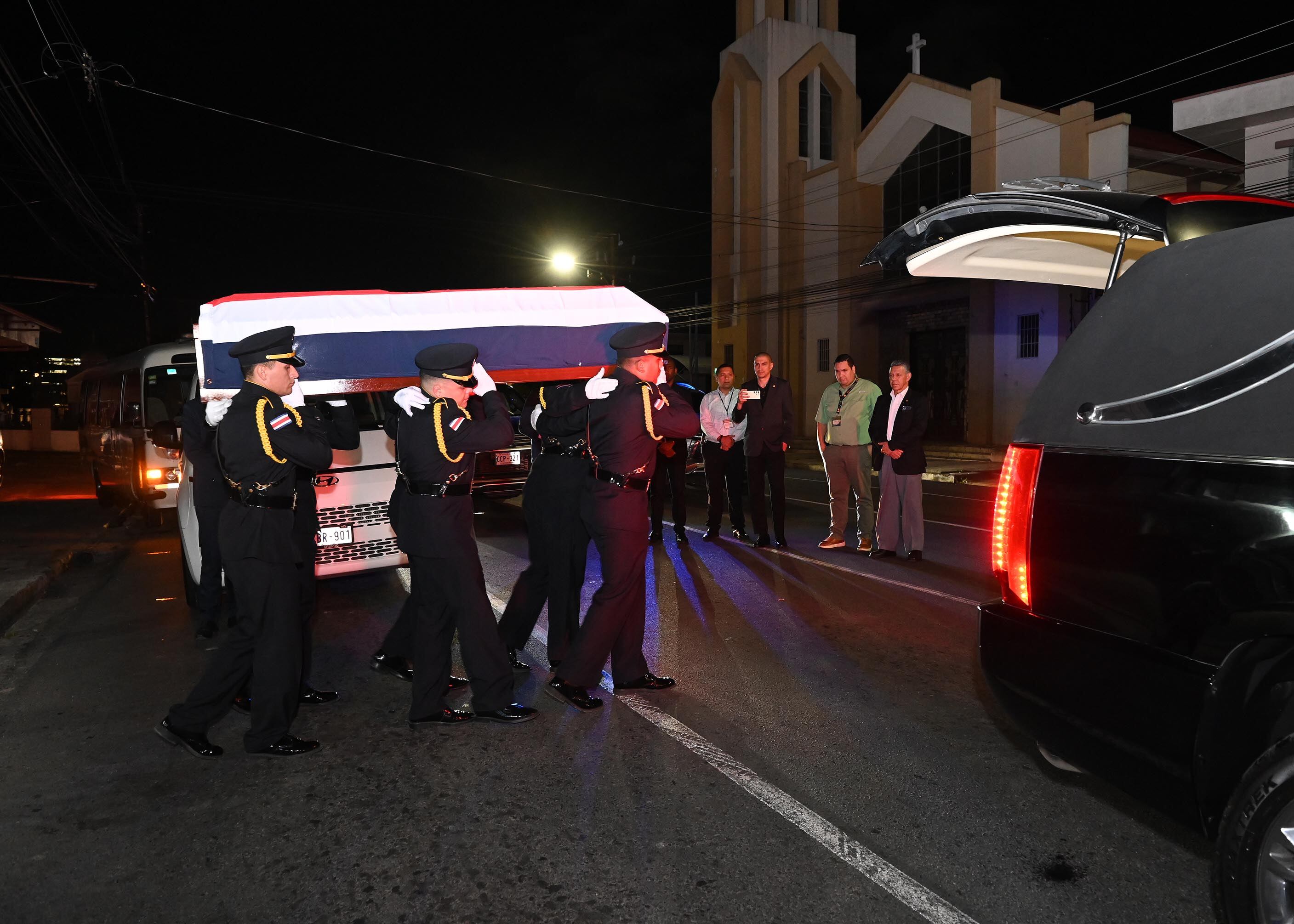 Policías vestidos de gala cargan un féretro y caminan hacia el carro funerario. La escena se desarrolla de noche.