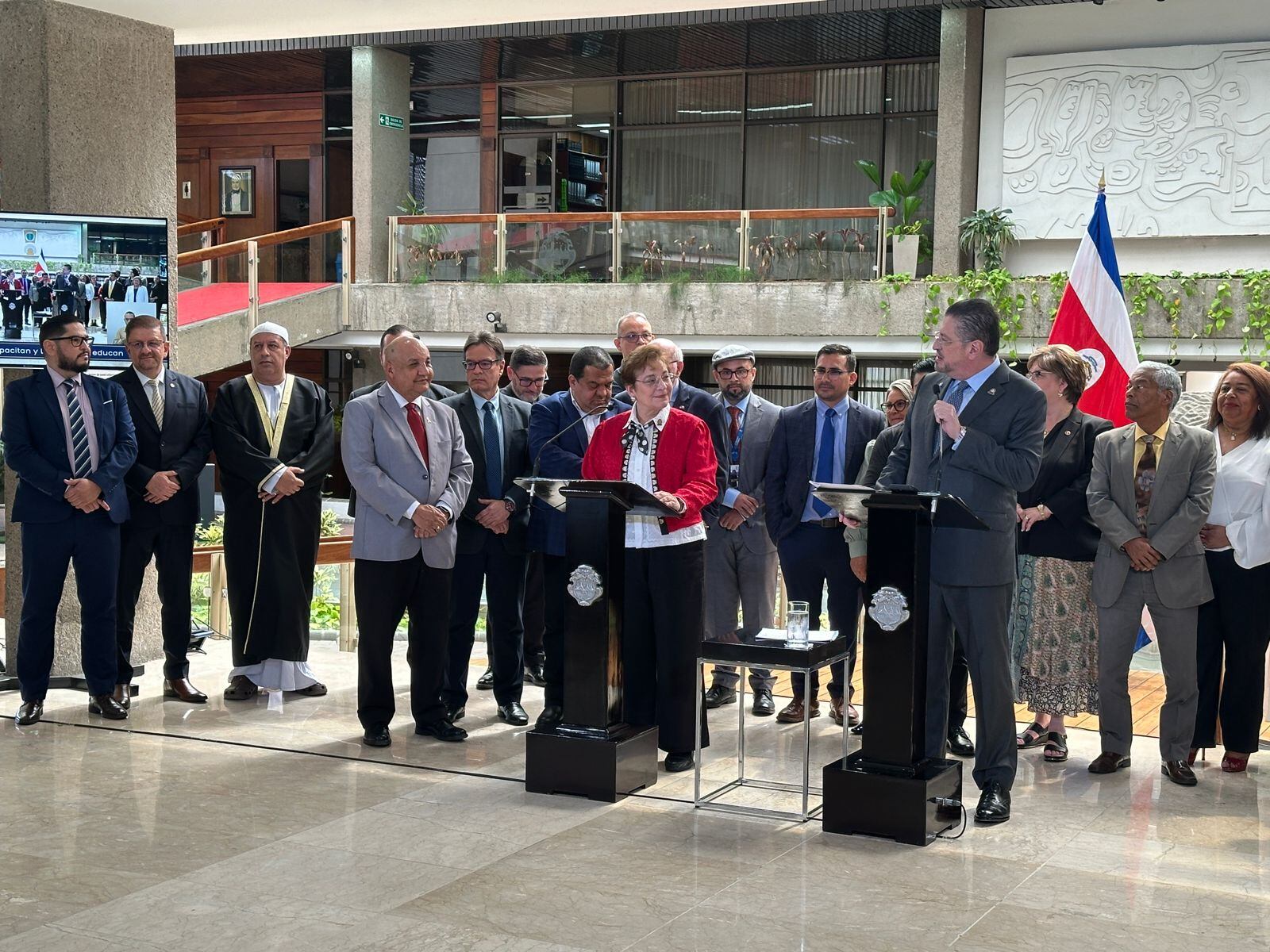 Se observa un grupo de personas que parecen respaldar a una mujer y a un hombre que se dirigen a los demás, desde un podio.