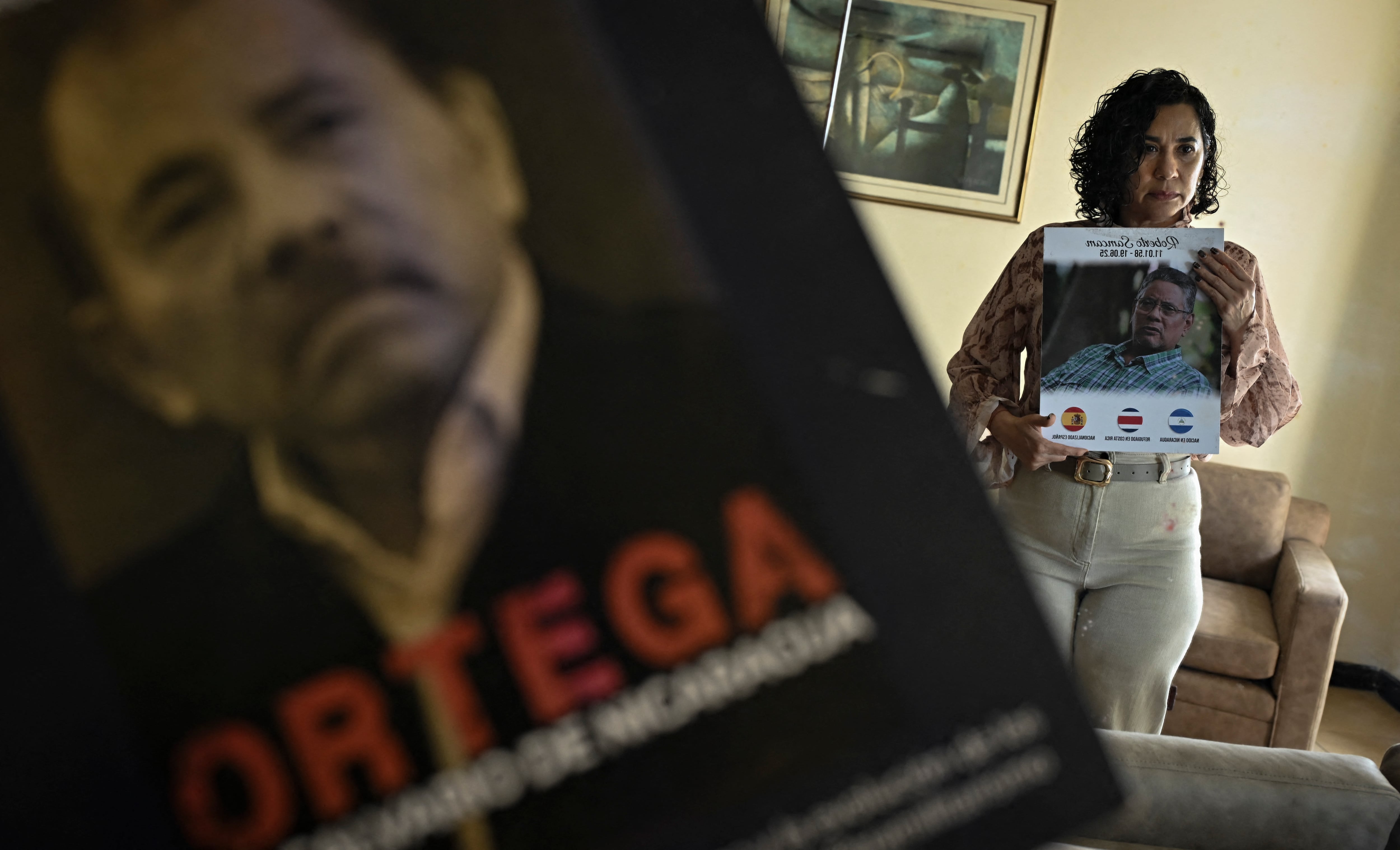 Claudia Vargas, viuda del exmayor retirado del Ejército de Nicaragua Roberto Samcam, posa junto a un retrato de su esposo y uno de los libros que escribió contra el gobierno de Daniel Ortega, en su residencia en San José. Samcam, exiliado en Costa Rica desde 2018, fue asesinado a balazos fuera de su vivienda en San José. (Foto por MARTIN BERNETTI / AFP)