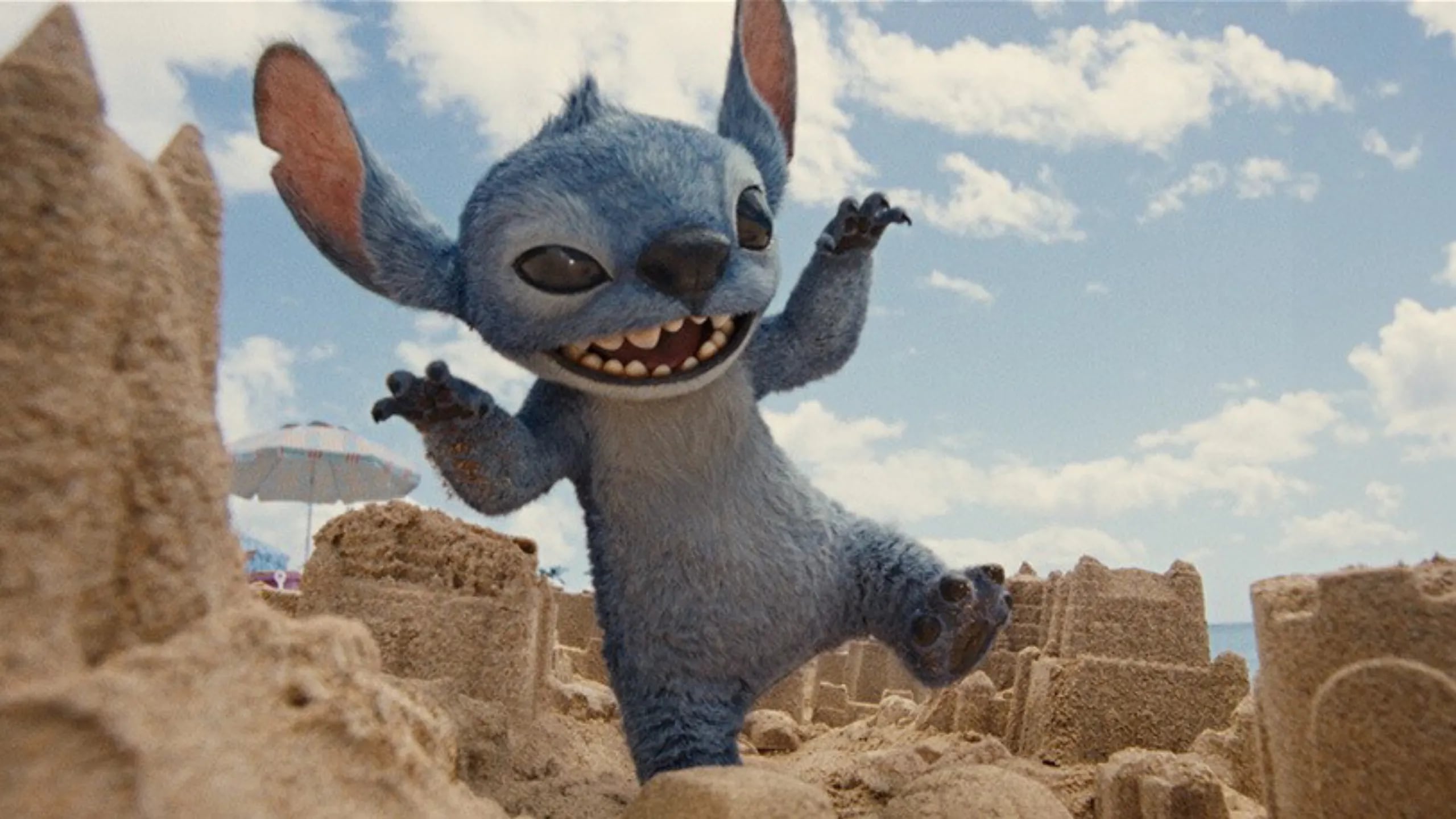 Stitch, personaje animado azul de la película Lilo & Stitch, aparece caminando alegremente sobre castillos de arena en una playa soleada, en una escena de la nueva versión live-action que se estrenará en Costa Rica en mayo de 2025.