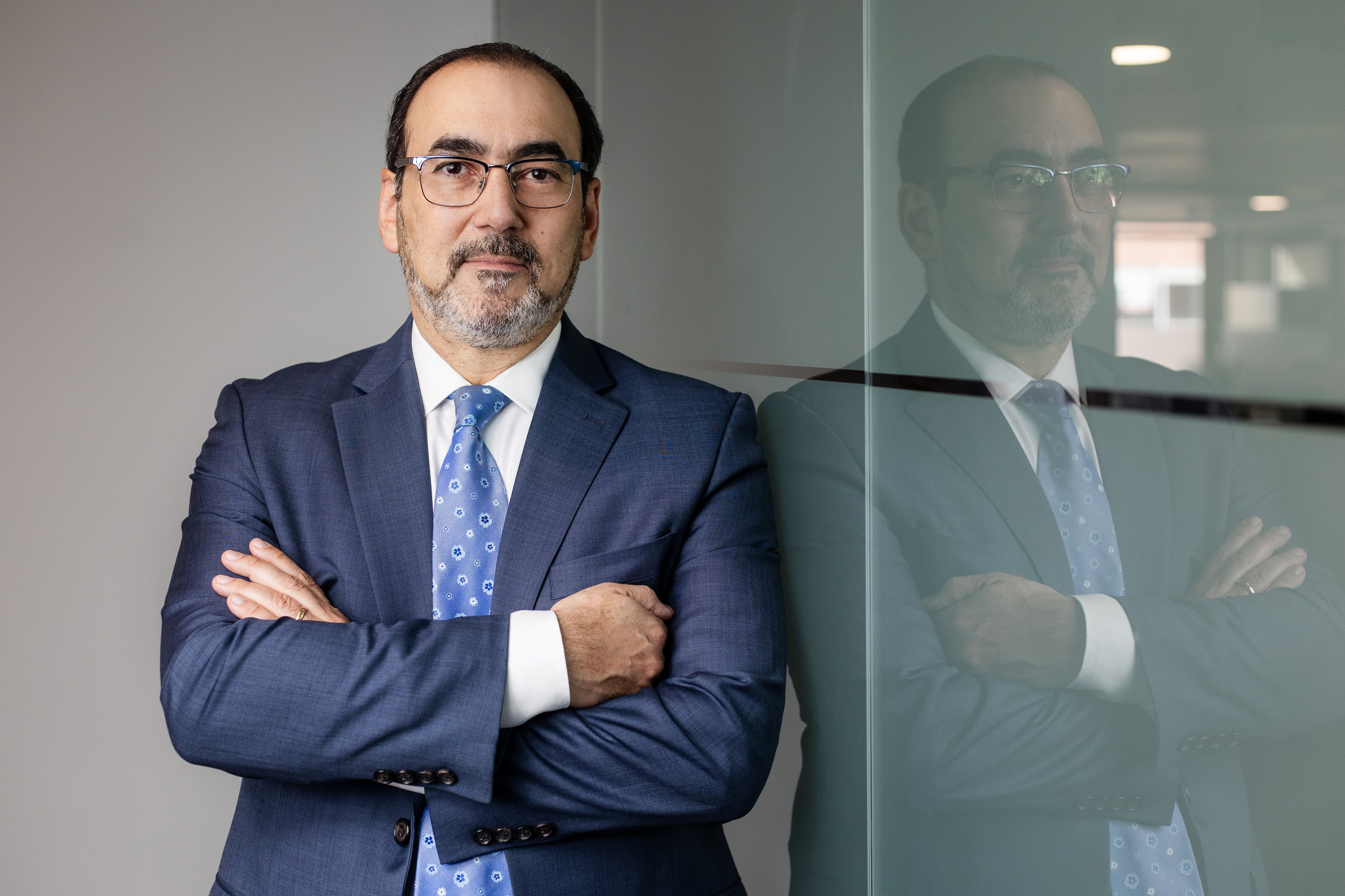 Sergio Díaz-Granados, presidente de CAF, posa con los brazos cruzados en un entorno corporativo. Su reflejo en un vidrio enfatiza su presencia en el sector financiero y su liderazgo en América Latina.