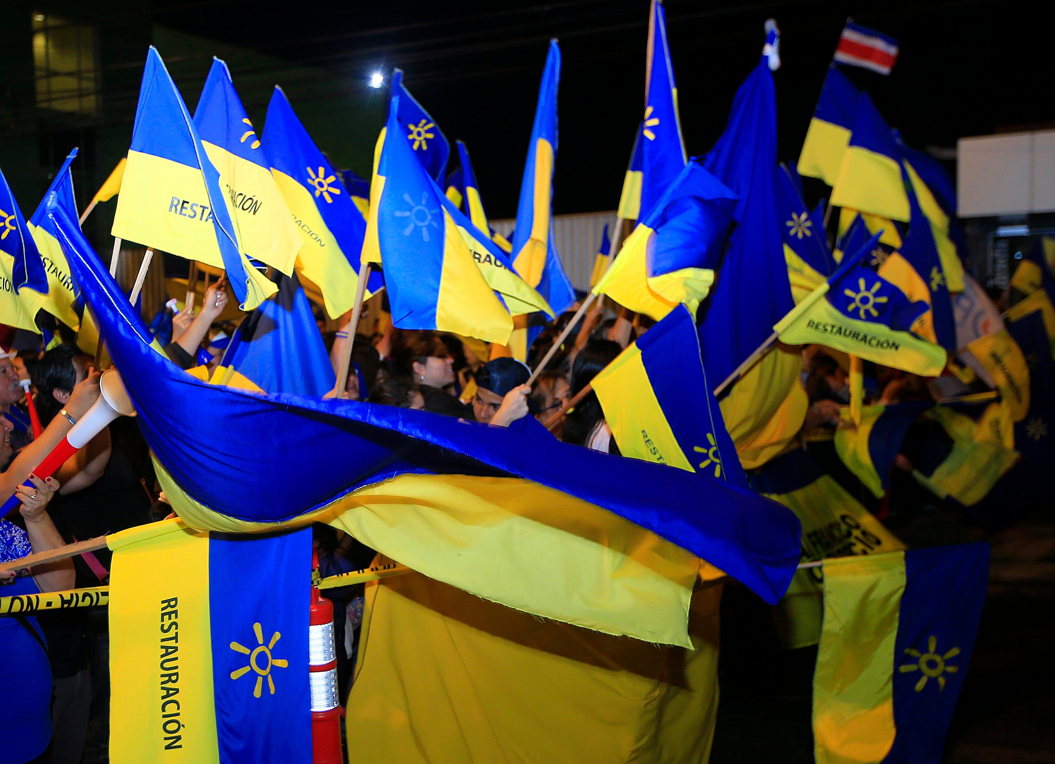 Banderas amarillas y azules del Partido Restauración Nacional ondeando durante un evento político nocturno, rodeadas de simpatizantes.