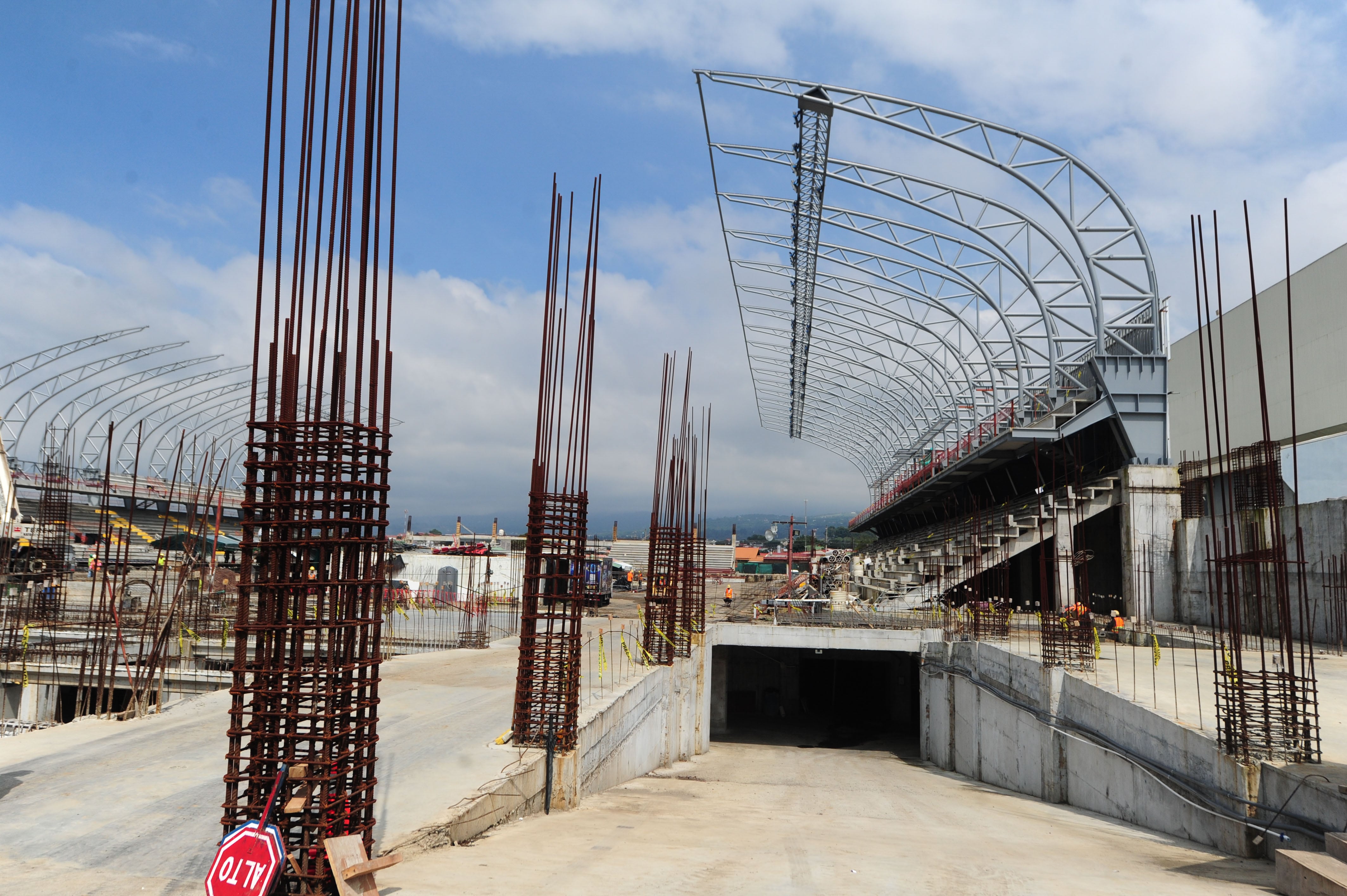 Construcción del Estadio Eladio Rosabal Cordero con columnas de acero, graderías en progreso y un cielo despejado al fondo.
