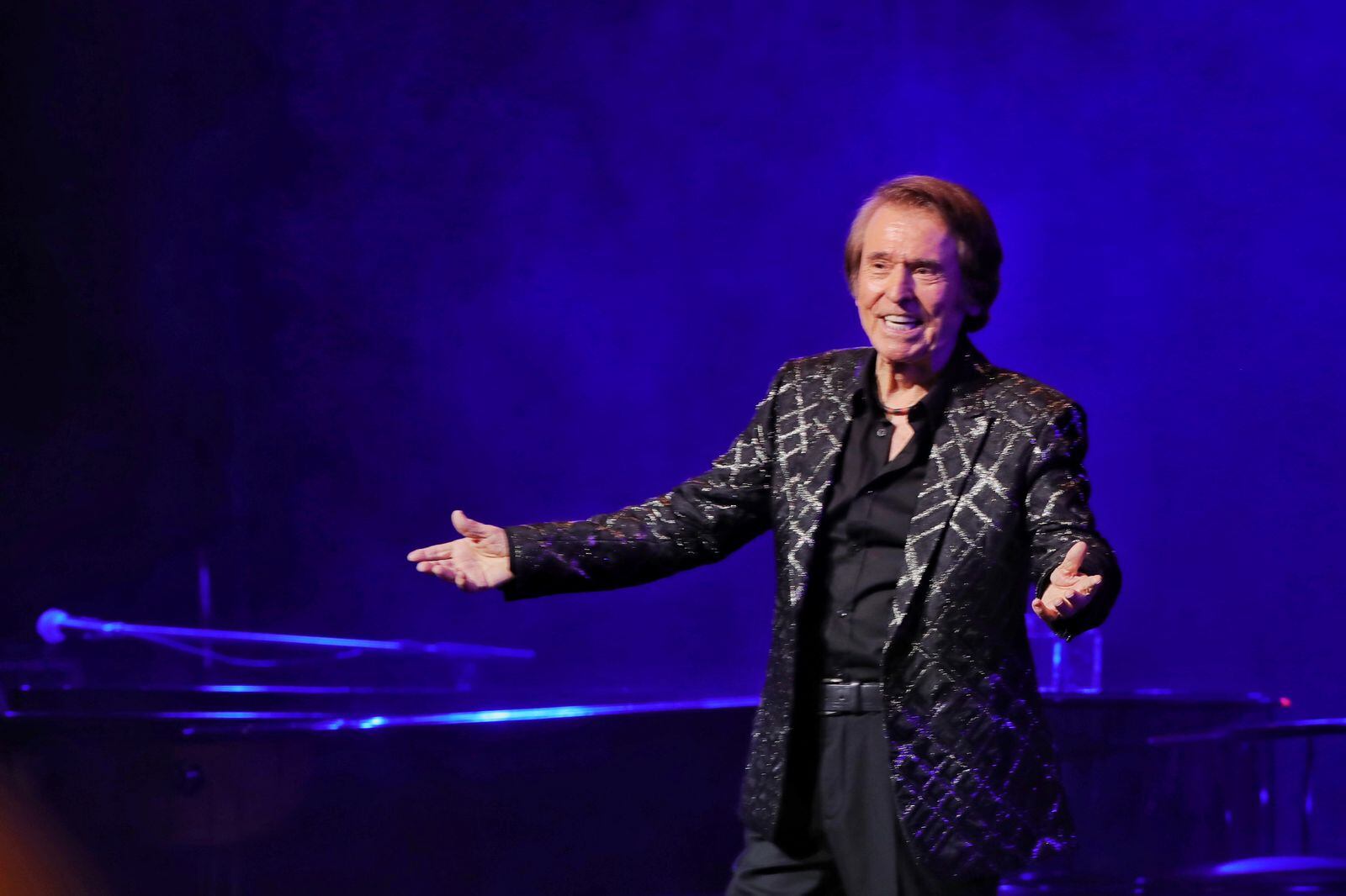 Imagen del cantante español Raphael durante uno de sus últimos conciertos en Costa Rica. Viste un traje de tono oscuro con camisa de color negro. Está con las manos extendidas.