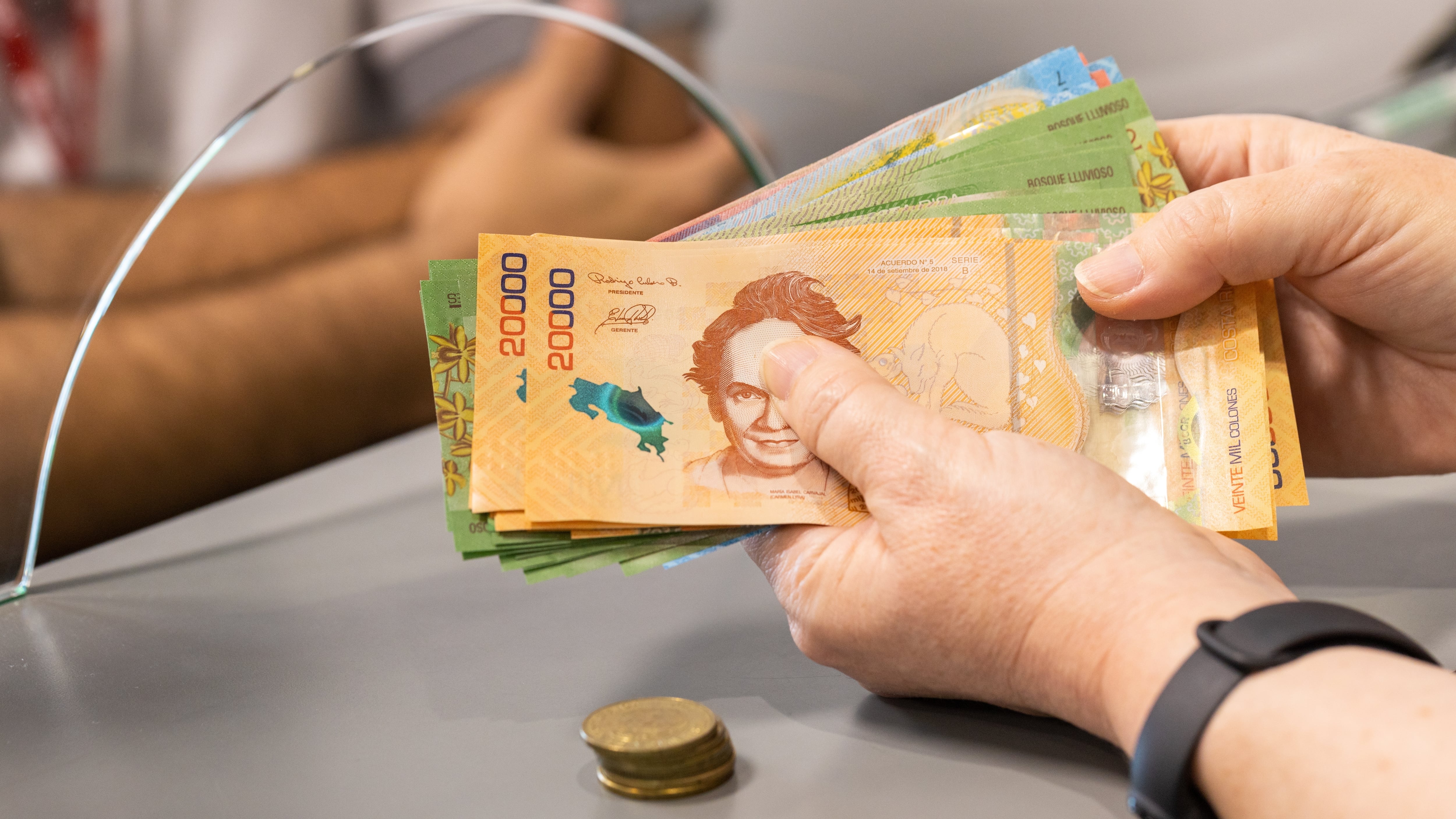 Manos de una persona contando billetes de 20.000 colones en un banco en Costa Rica, con un empleado detrás de un vidrio de atención.