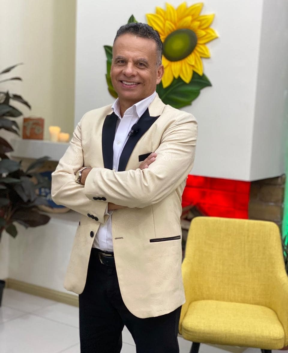 Imagen del periodista Giovanni Calderón posando para la foto, viste un saco de color rosa, camisa blanca y pantalón negro. Sonríe y tiene los brazos cruzados sobre su pecho.