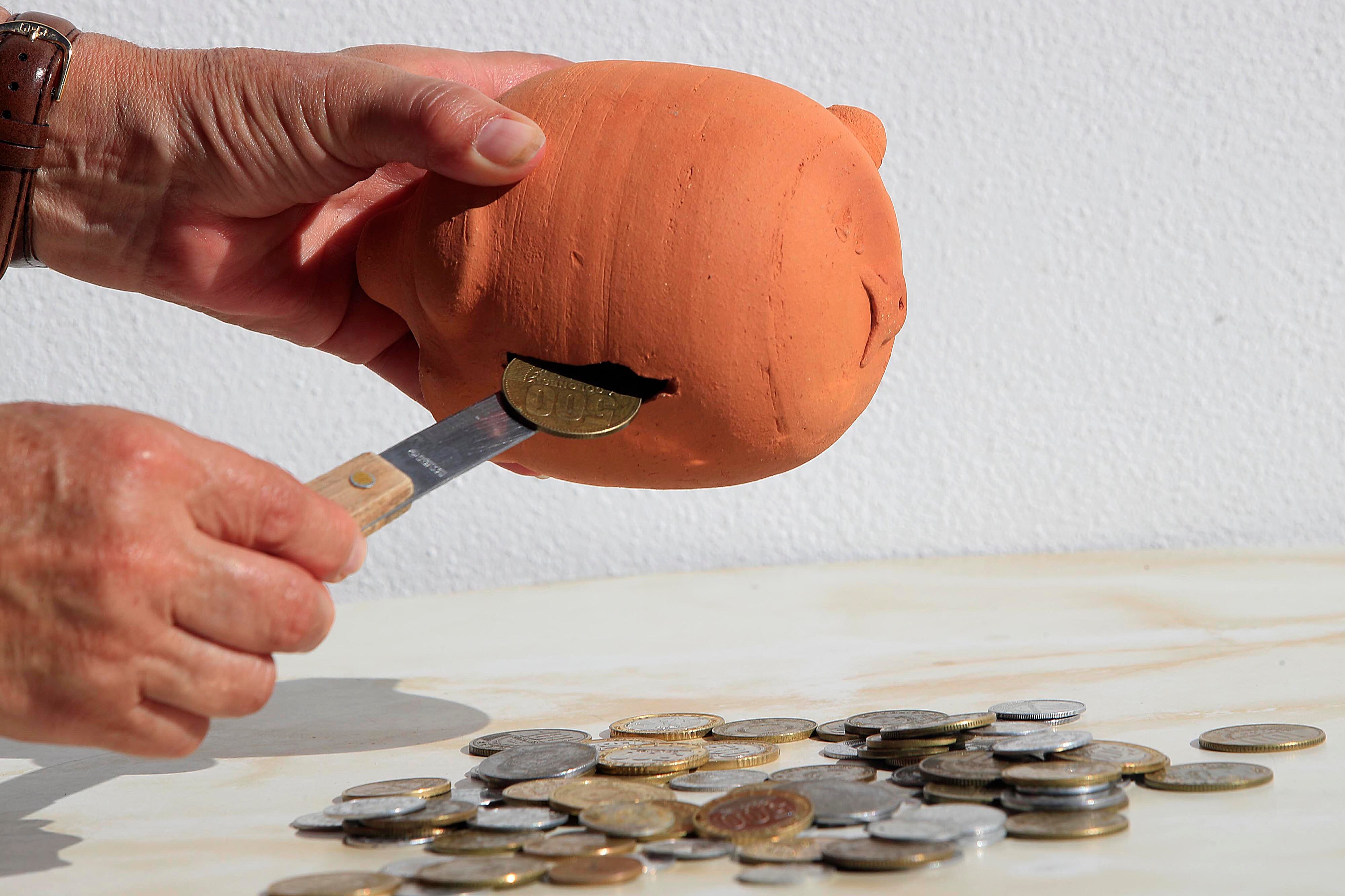 Manos sacando monedas de una alcancía de barro con un cuchillo, junto a un montón de monedas en una superficie, representando los ajustes financieros de la cuesta de enero.
