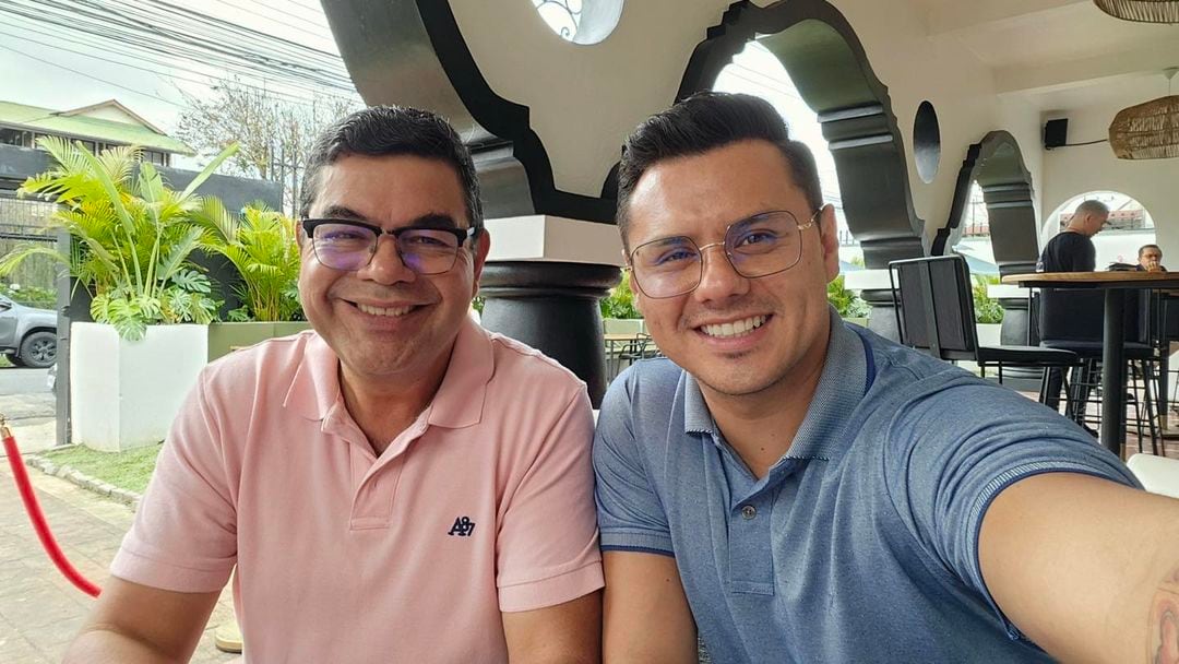 El periodista Marcelo Castro y su pareja el también comunicador Johnny López. Están sentado junto al otro, Marcelo usa camisa rosada y Johnny una azul. Ambos sonríen.