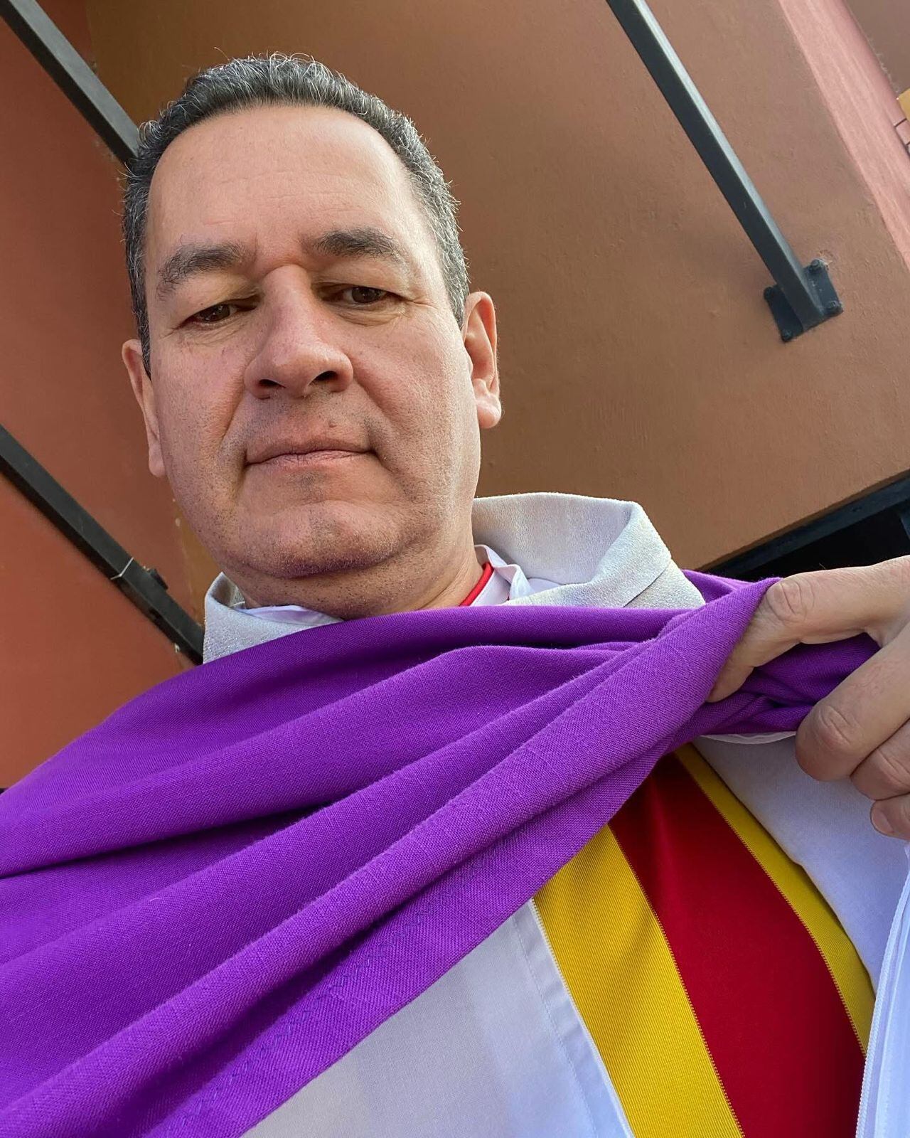 El sacerdote conocido como el padre Sejo se abre sus prendas sacerdotales y muestra que por debajo viste una camiseta de franjas verticales en rojo y amarillo del Club Sport Herediano.