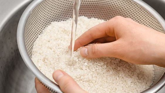 Arroz crudo en un recipiente, le cae un chorro de agua y unas manos lavan los granos.