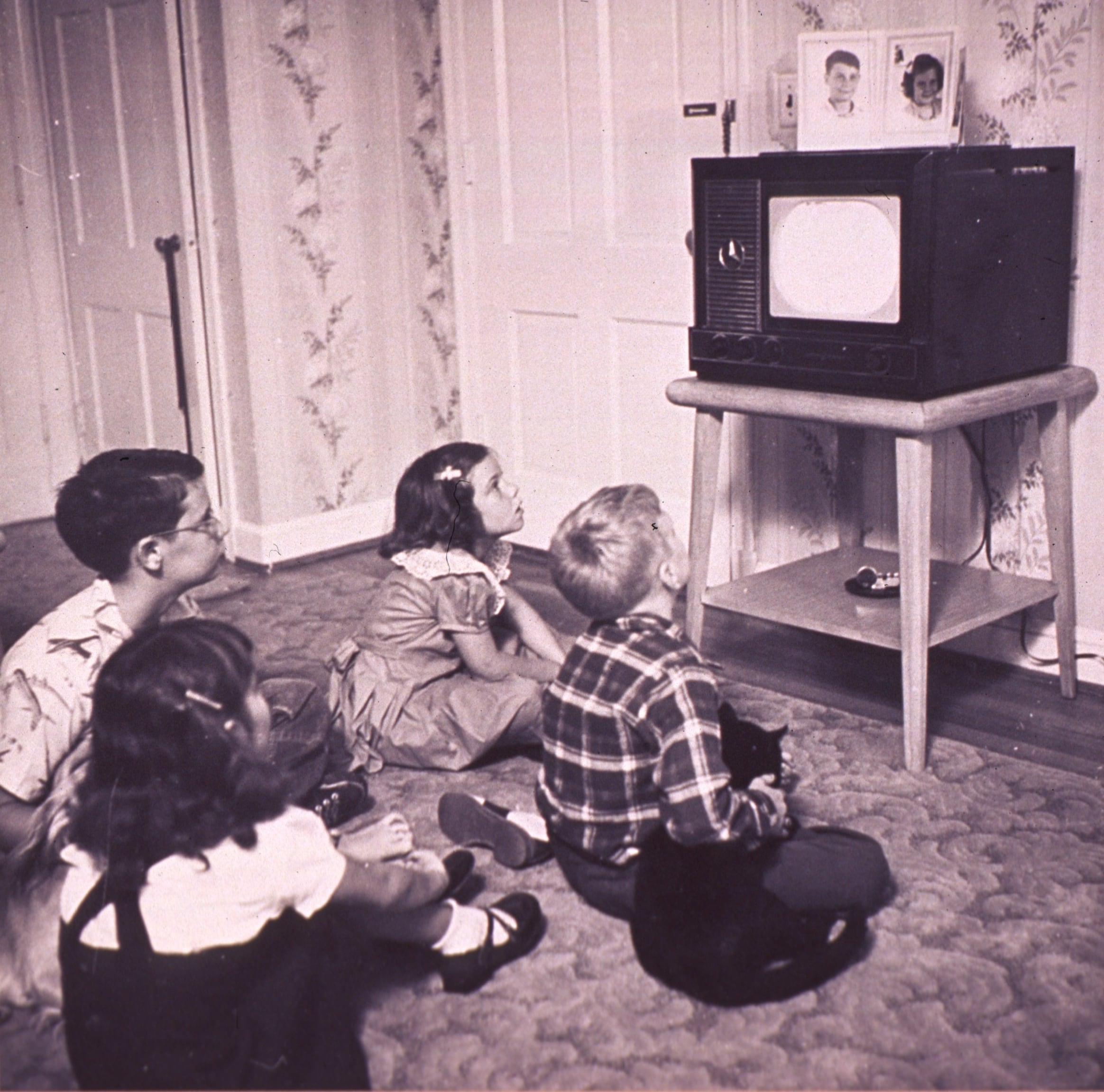 Cuatro niños sentados en el suelo, viendo televisión en un aparato clásico, vintage en blanco y negro