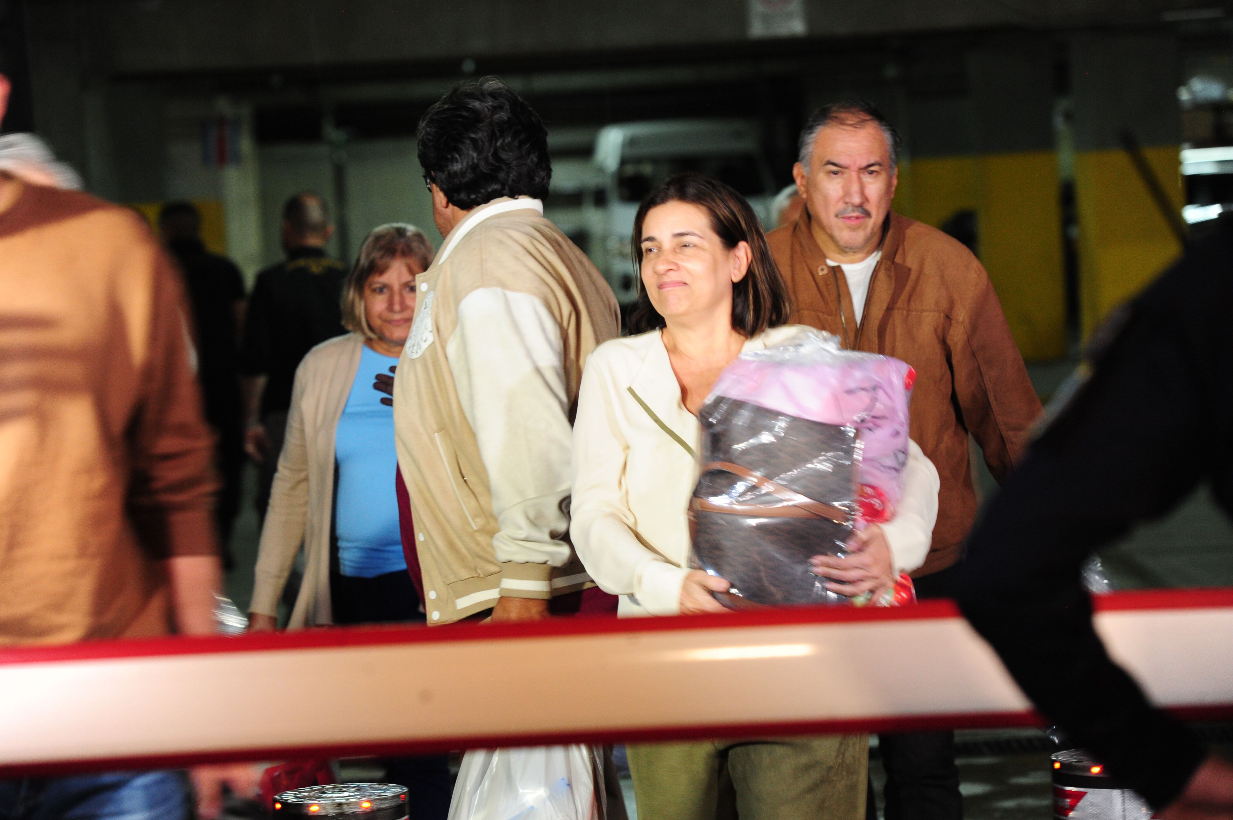 Grupo de cuatro personas. Primer plano, una mujere de mediana edad, traje claro, carga una bolsa con objetos personales en sus brazos.