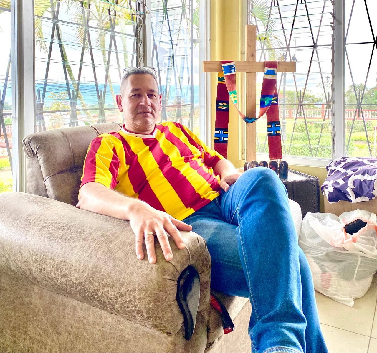 Sacerdote sentado en la sala de la casa cural de Mata de Plátano, vistiendo una camiseta del Club Sport Herediano.