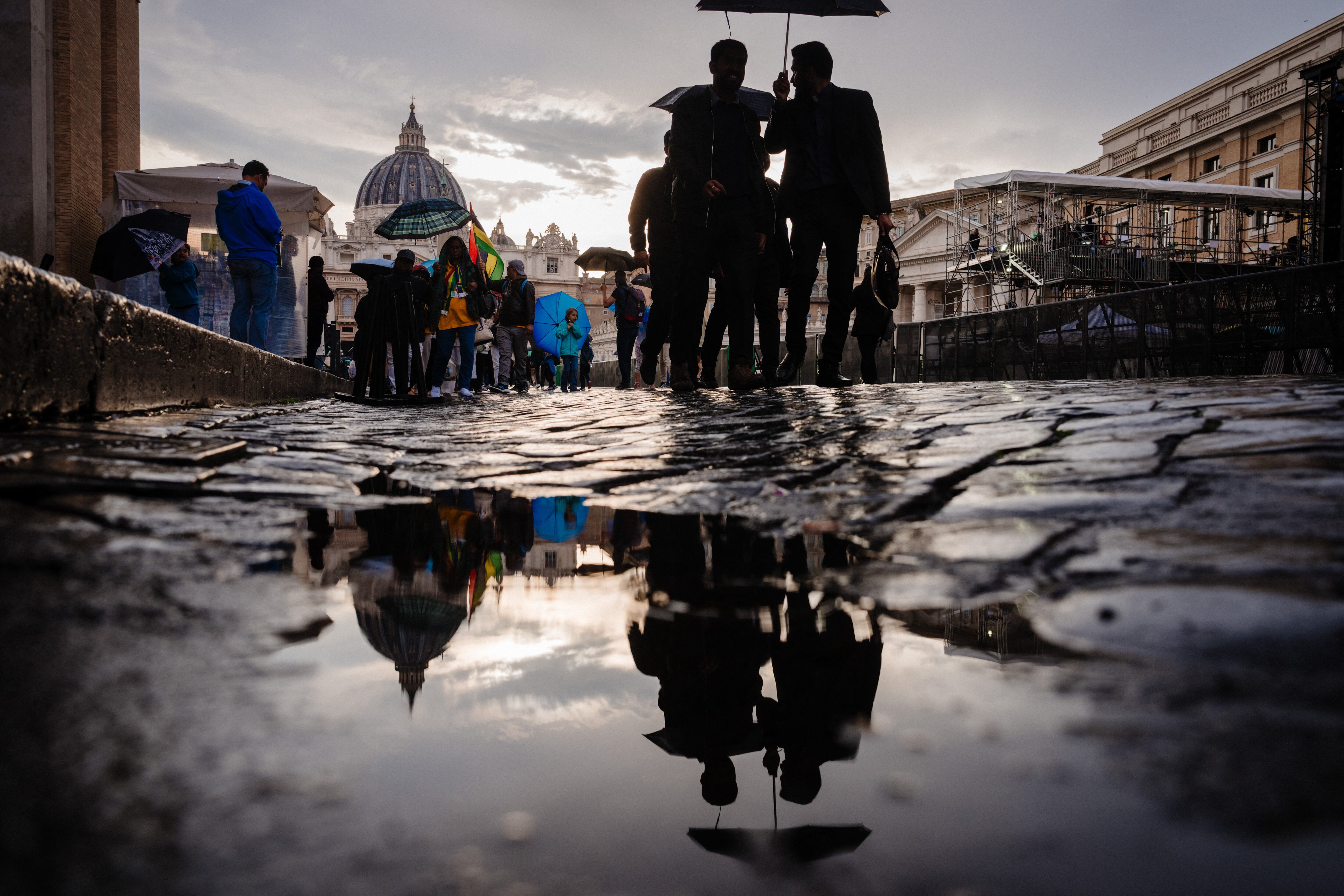 Peatones se reflejan en un charco con la basílica de San Pedro al fondo, cerca del Vaticano, en Roma, el 28 de abril de 2025. (Foto por Dimitar DILKOFF / AFP)