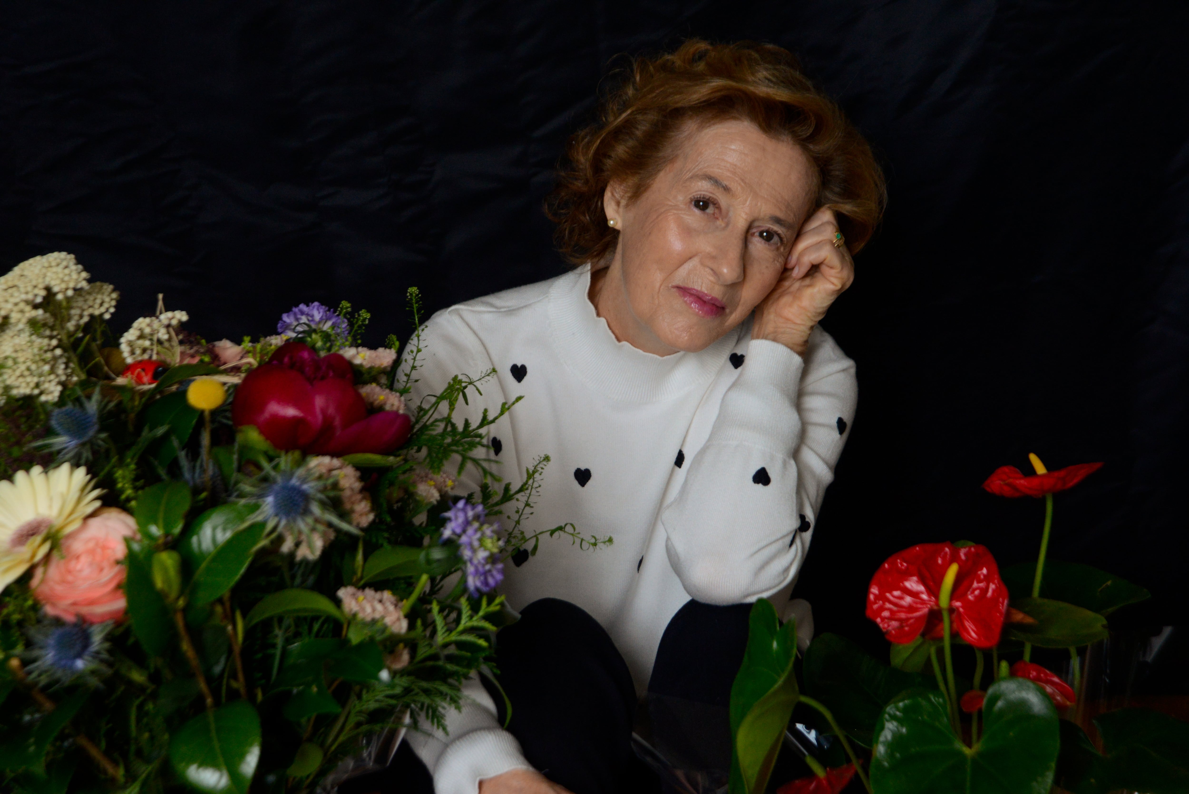 Escritora española Julia Navarro posando para la cámara. Hay flores delante de ella, tiene una mano en la cara, usa blusa blanca.