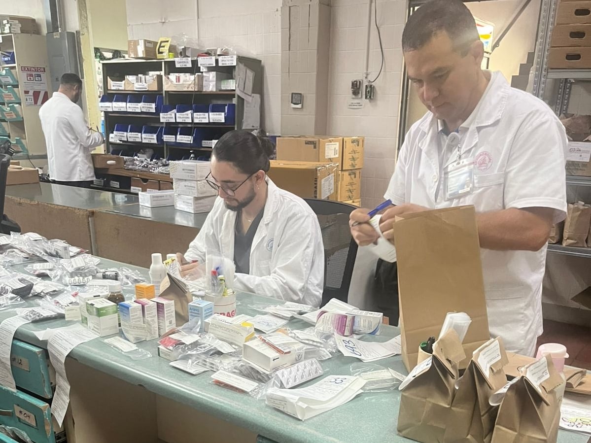 Dos hombres jóvenes vestidos con gabacha blanca de manga corta empacan medicamentos de un hospital.