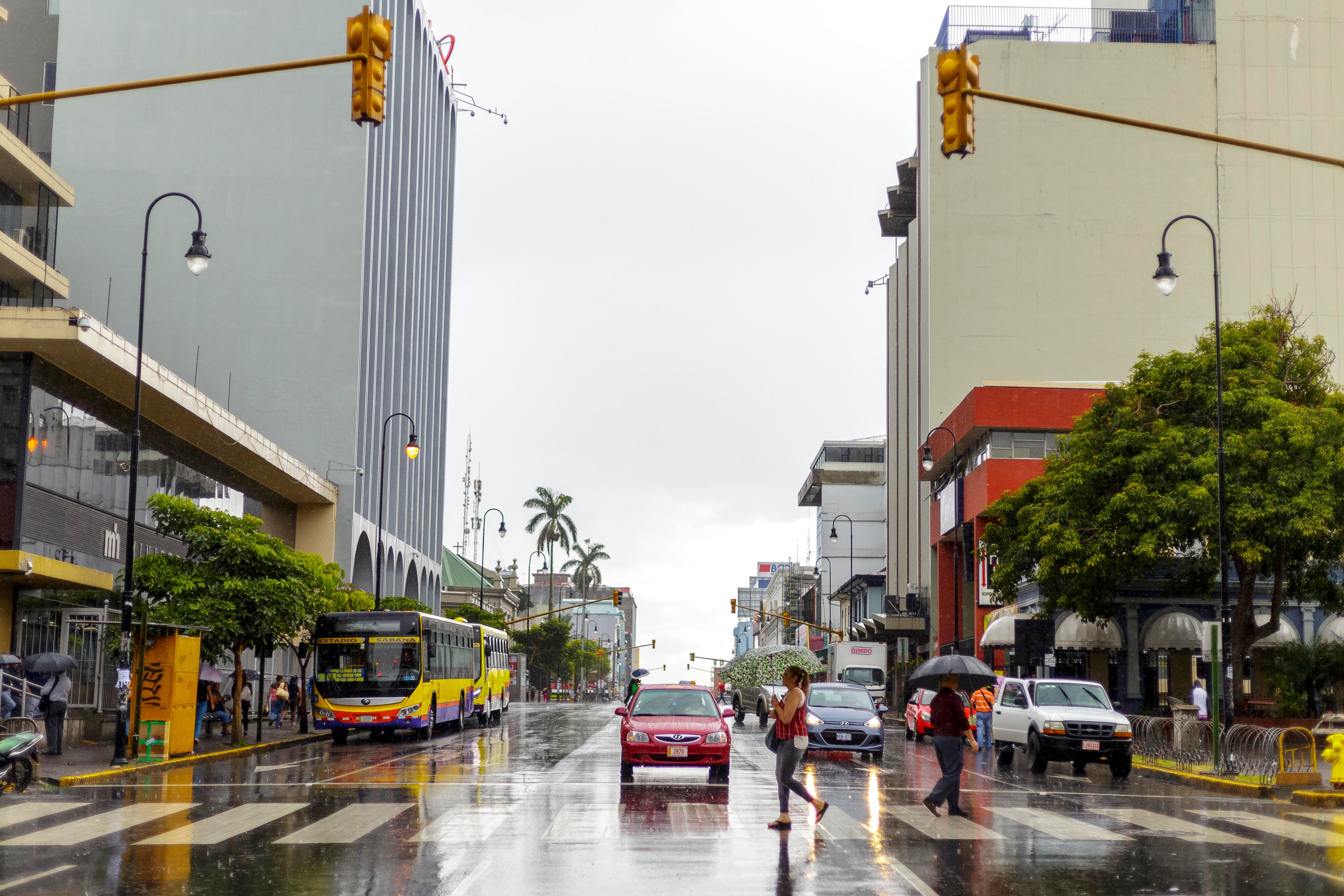 Calle del casco central de San José con vehículos, un autobús y peatones cruzando en medio de un día lluvioso, destacando edificios y semáforos encendidos.