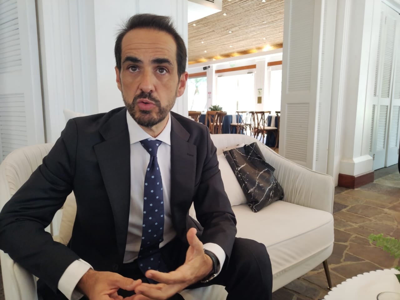 Francisco Javier Urra, representante del BID en Costa Rica, viste traje oscuro y corbata azul mientras conversa en una entrevista en un espacio con muebles blancos.