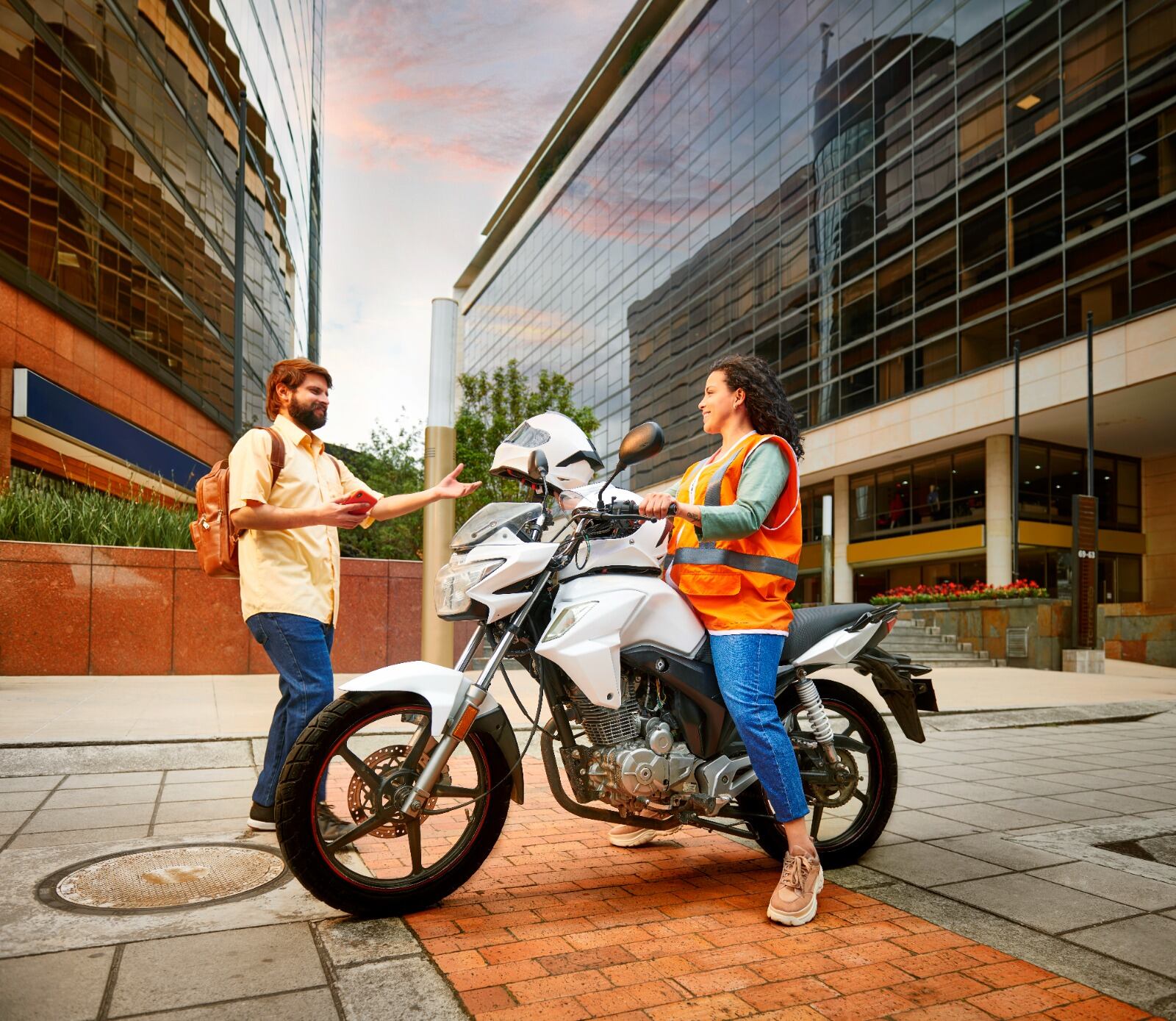 Un hombre con mochila habla con una conductora de DiDi Moto, quien está apoyada en su motocicleta y usa un chaleco reflectante. La imagen muestra el nuevo servicio de transporte en motocicleta que la empresa lanzó en Costa Rica.