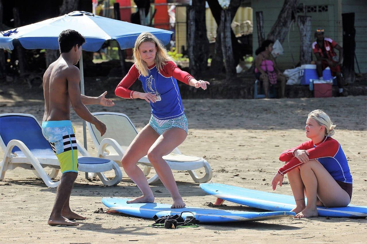 Turista recibe instrucciones de surf en la arena de una playa costarricense, mientras otra persona observa sentada en una tabla de surf y el instructor guía.