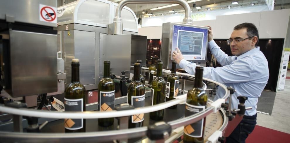 Hombre monitoreando el funcionamiento de una línea automatizada de producción de botellas de vino en una planta manufacturera, con pantalla de control y botellas en proceso.