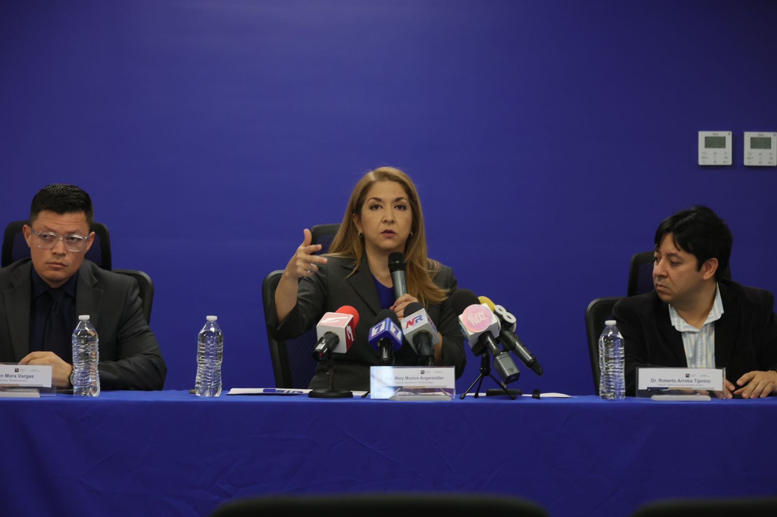Tres personas: dos hombres y una mujer al centro, sentados a la orilla de una mesa, con mantel azul, fondo azul, con micrófonos al frente. Conferencia de prensa.