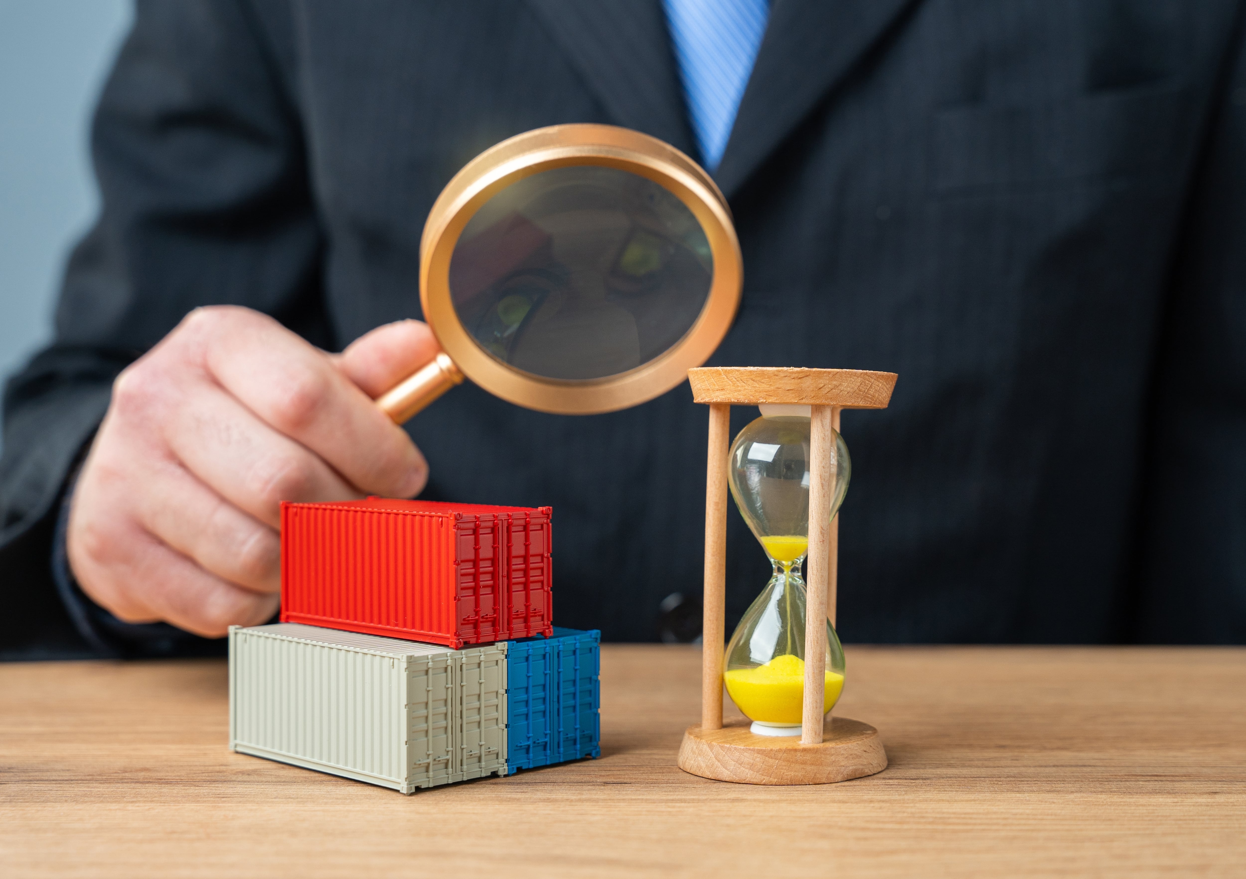 Mano con lupa inspecciona contenedores de carga junto a un reloj de arena, símbolo de barreras comerciales, regulaciones e impacto en el comercio entre Estados Unidos y Costa Rica