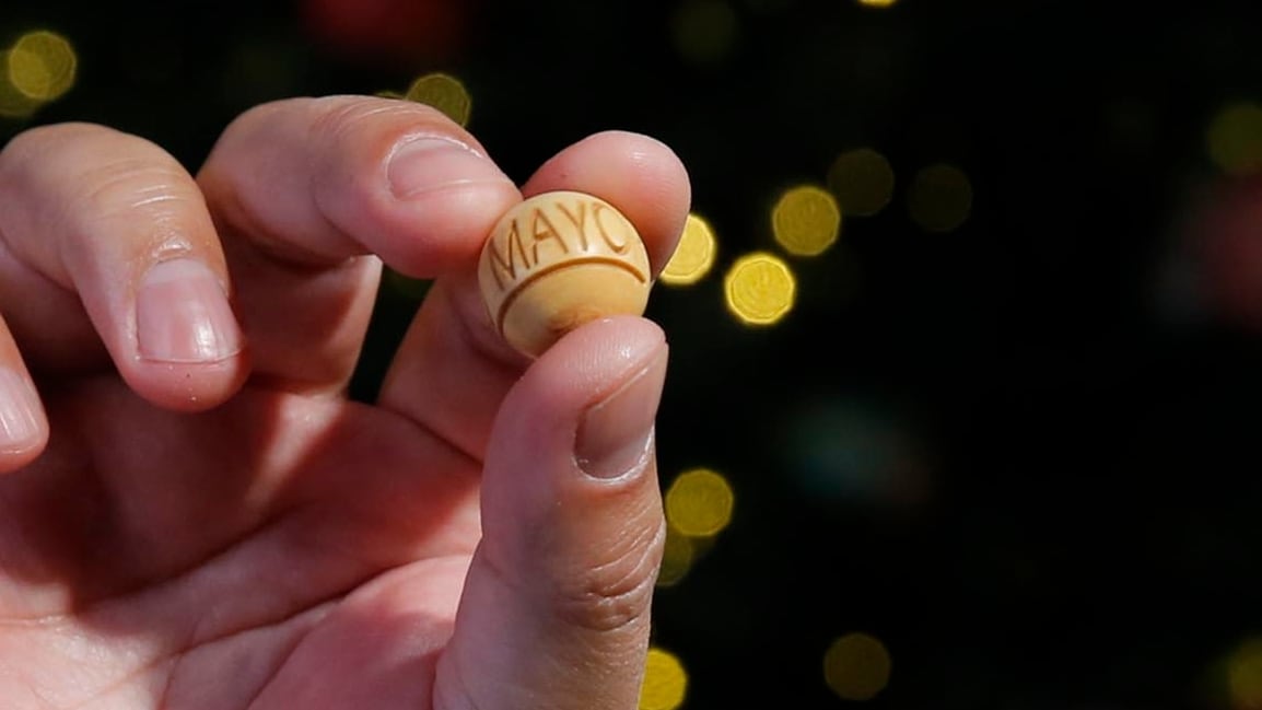 Mano sosteniendo una bola de sorteo del Gordo Navideño 2023 marcada con el mes de mayo, sobre un fondo decorativo con luces desenfocadas.