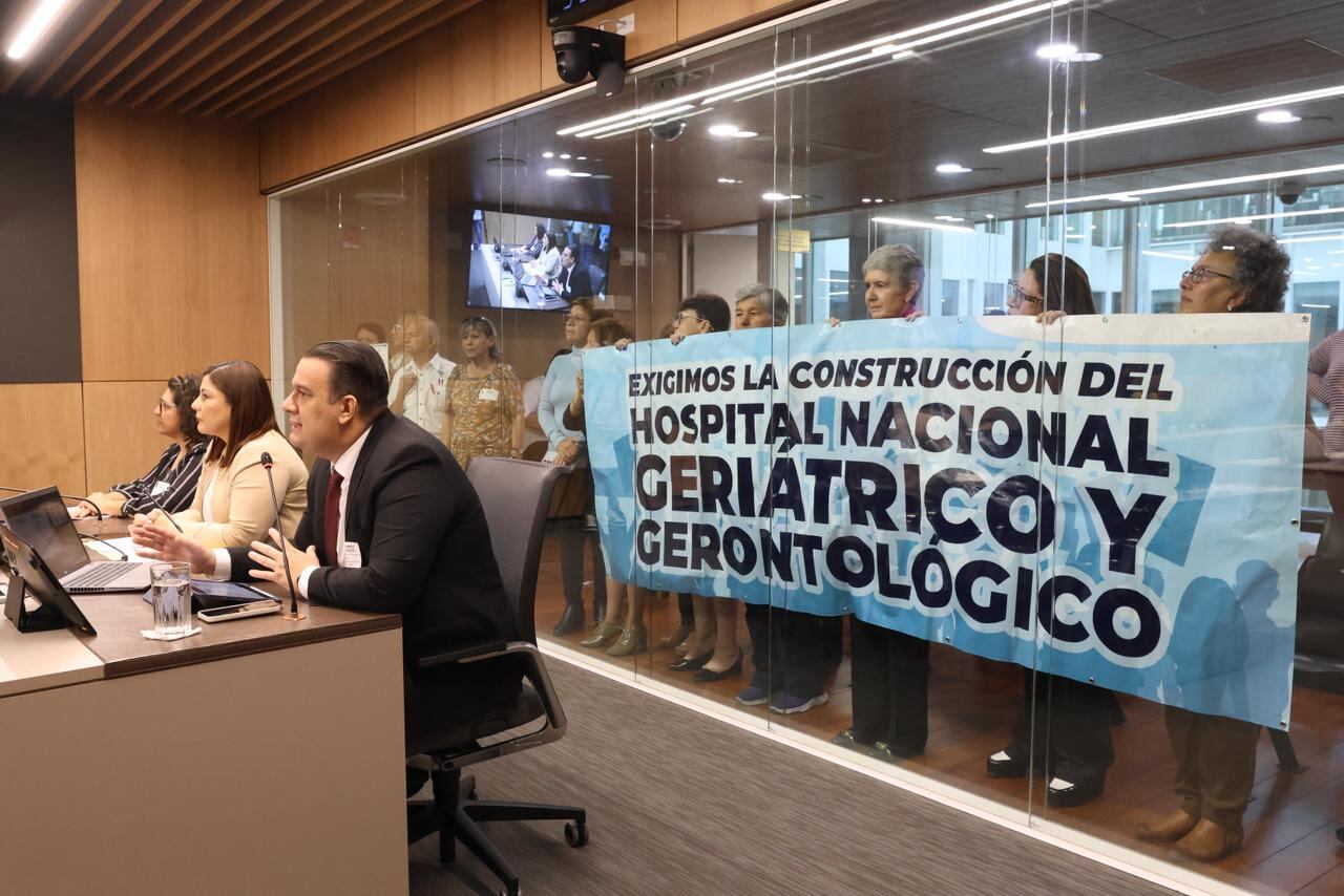 Se ve una sala de audencias, hay tres comparecientes y detrás de un vidrio un grupo del público sostiene una manta donde pide la construcción del hospital nacional de geriatría.