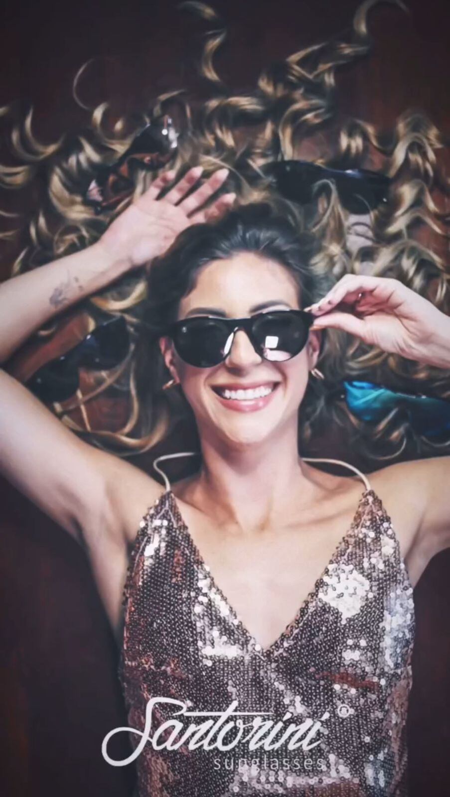 Plano cenital de una mujer acostada con su cabello suelto. Abajo se lee la leyenda Santorini Sunglasses