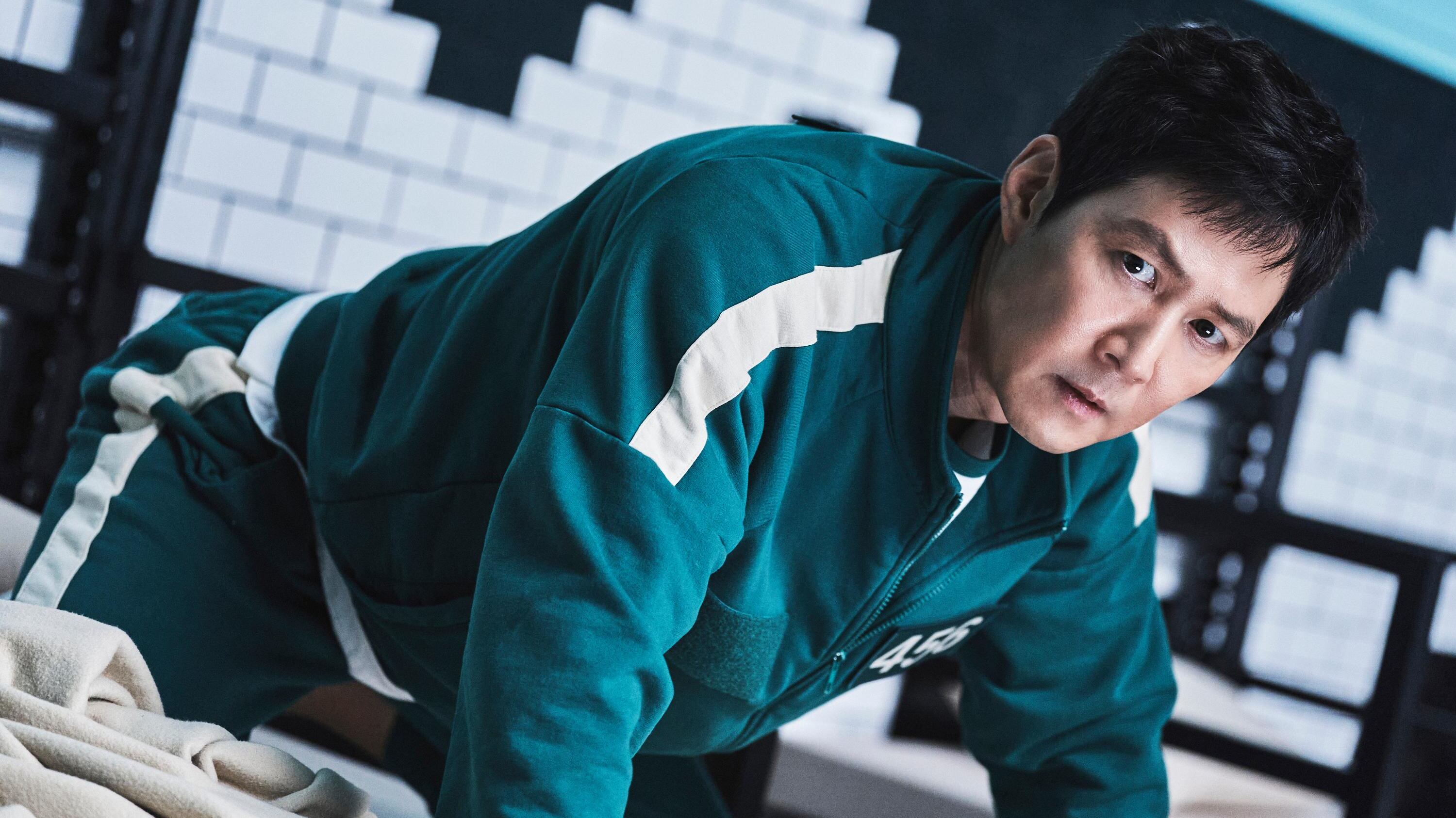 Escena de 'El Juego del Calamar' en la que Gi-hun aparece vestido como participante, hincado sobre una cama