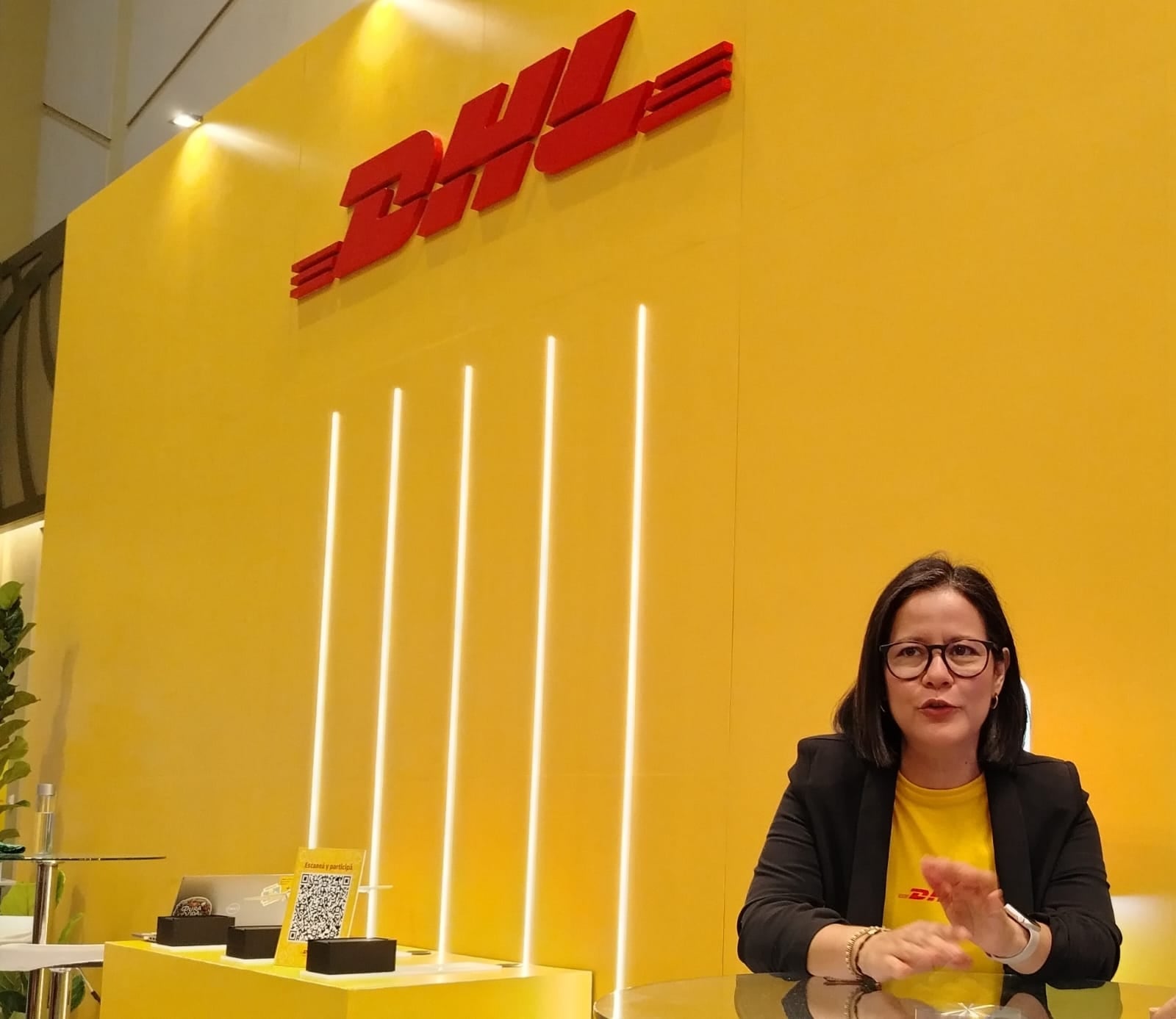 Claudia Ortiz, gerente país de DHL Costa Rica, participa en una entrevista durante una actividad empresarial. La ejecutiva aparece frente a un panel amarillo con el logotipo de DHL, mientras responde preguntas de periodistas en un espacio de exhibición corporativa.