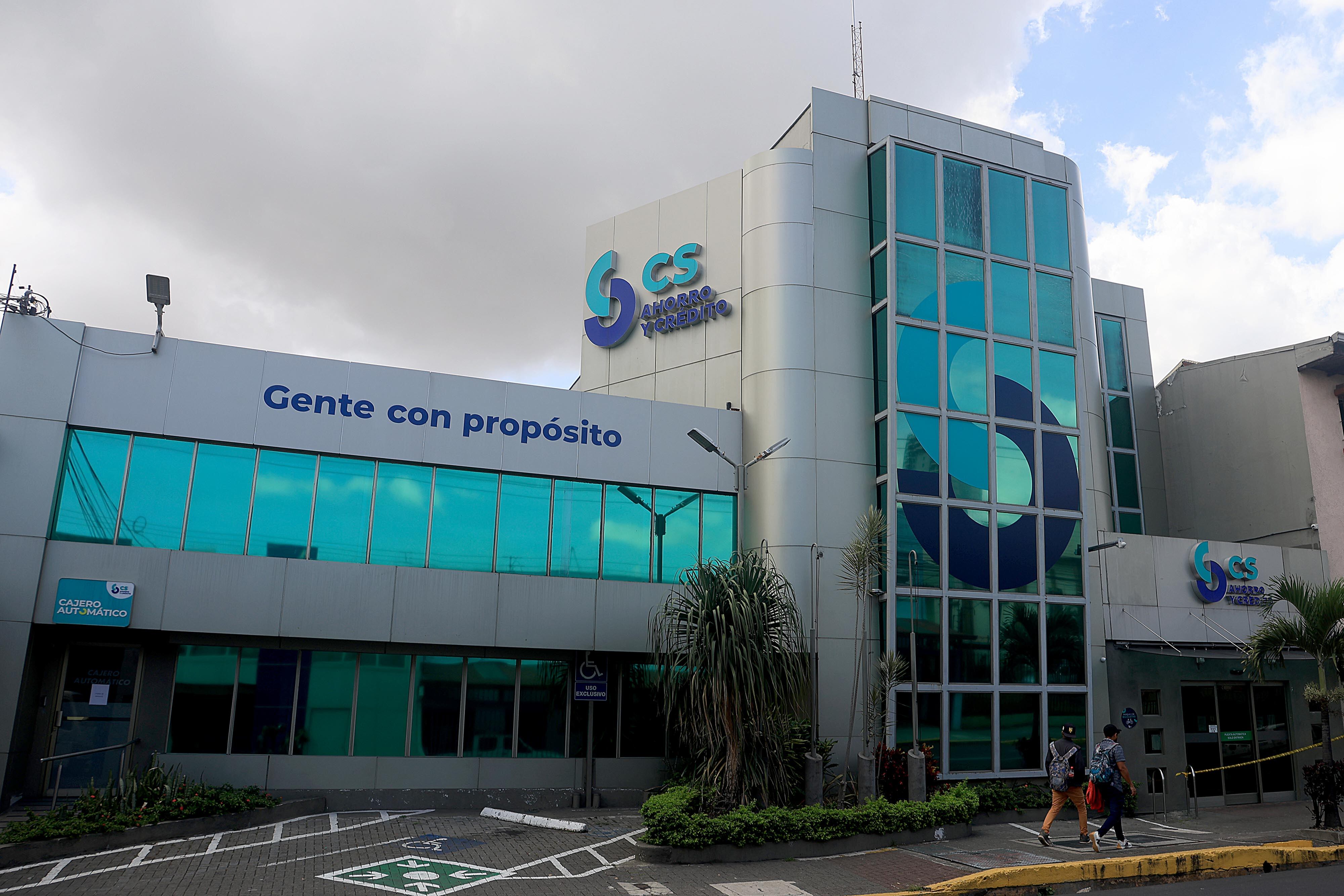 Fachada principal del edificio de Coopeservidores en Costa Rica, con letrero de "Gente con propósito", destacando ventanas de vidrio y un área de estacionamiento accesible en primer plano.