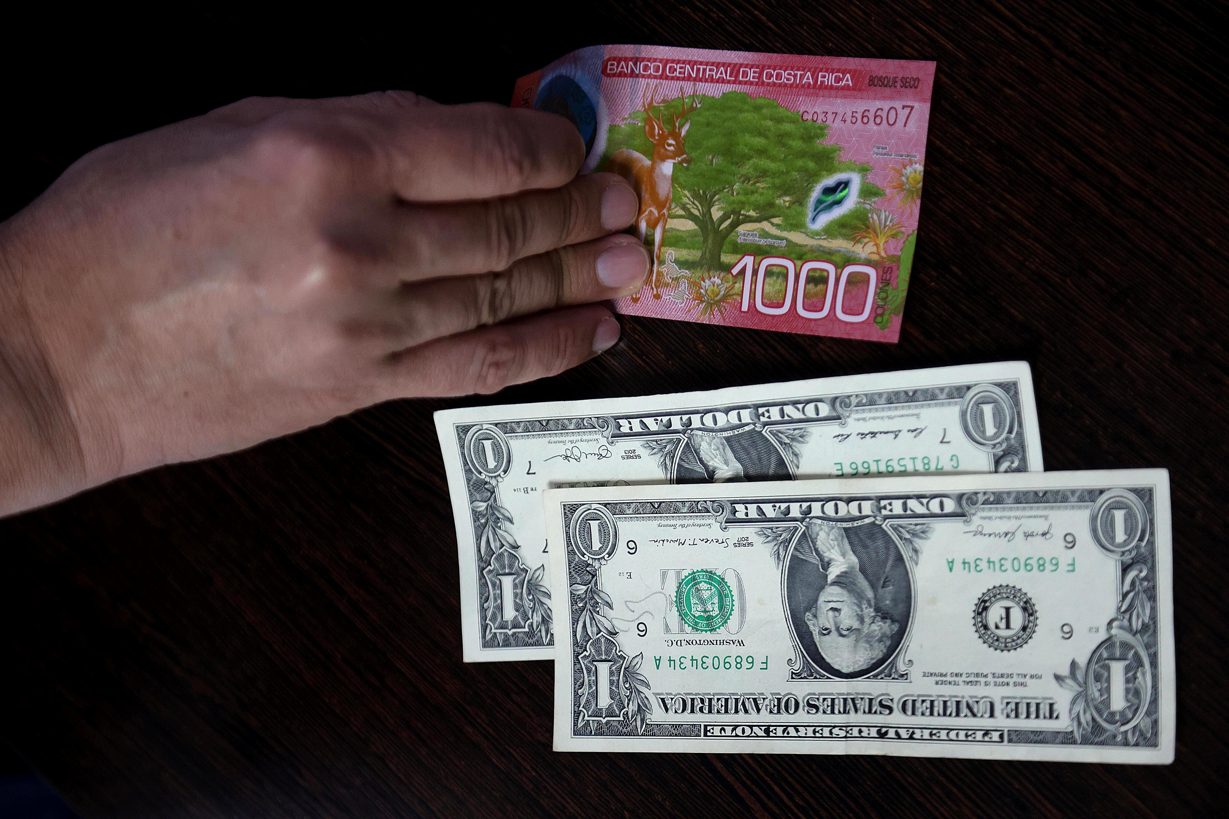 Una mano sostiene un billete de ¢1.000 colones junto a dos billetes de un dólar estadounidense sobre una mesa.