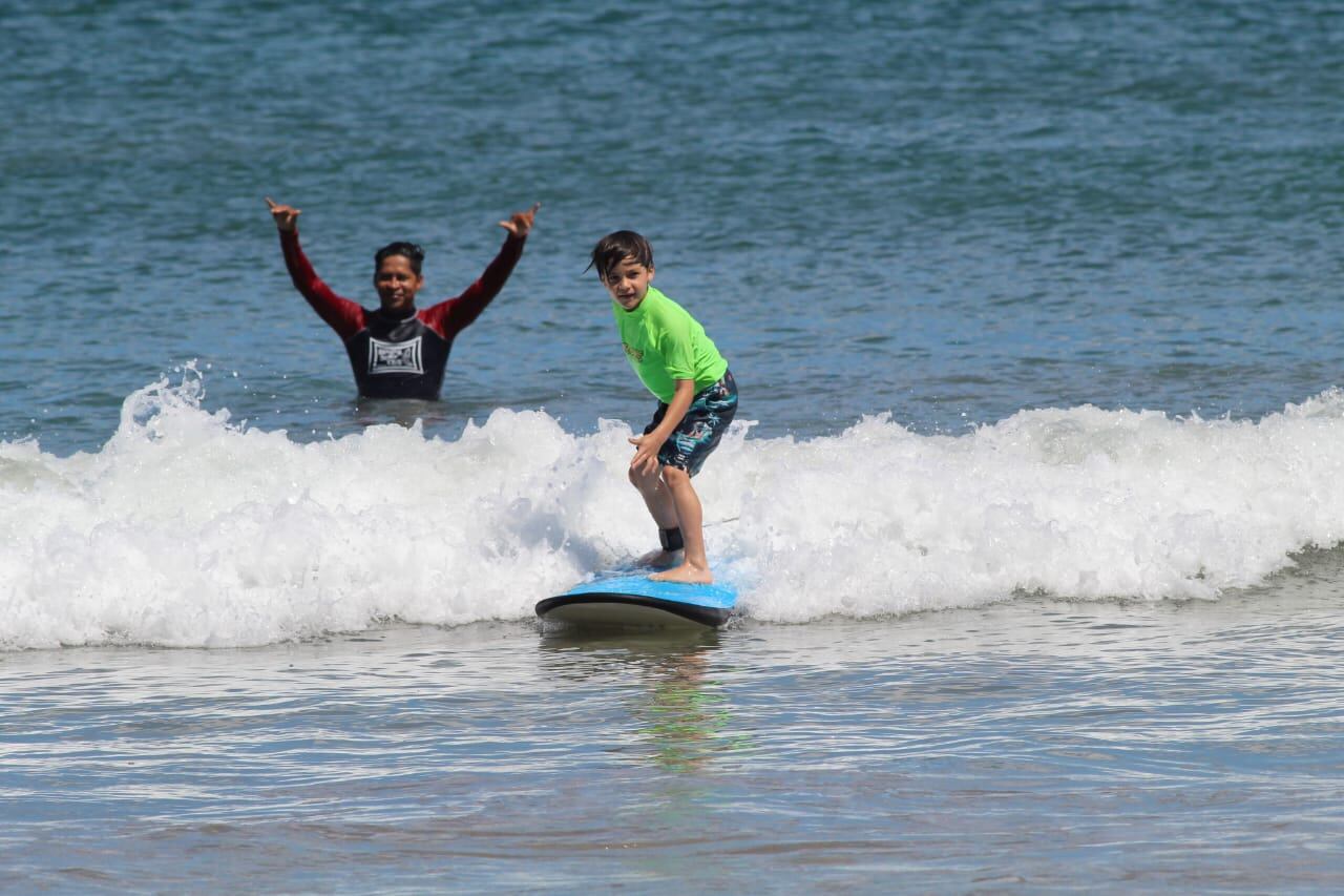 Niño de pie sobre una tabla de surf montando una pequeña ola, mientras un instructor lo guía desde el agua con señas en una playa de Costa Rica.
