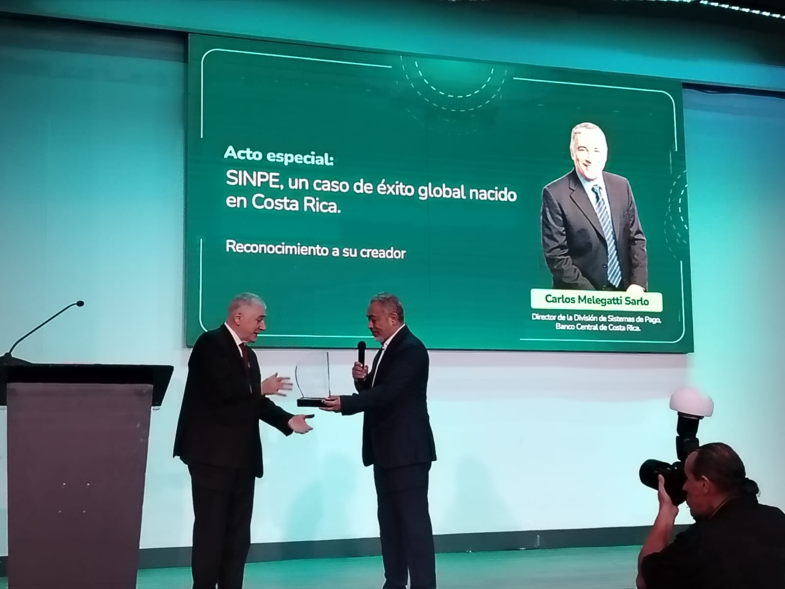 Dos hombres con traje participan en un acto de reconocimiento durante el Fintech Summit. Uno de ellos entrega una distinción al otro mientras, en la pantalla del escenario, se observa un mensaje sobre Sinpe y su desarrollo como sistema de pagos digitales en Costa Rica.