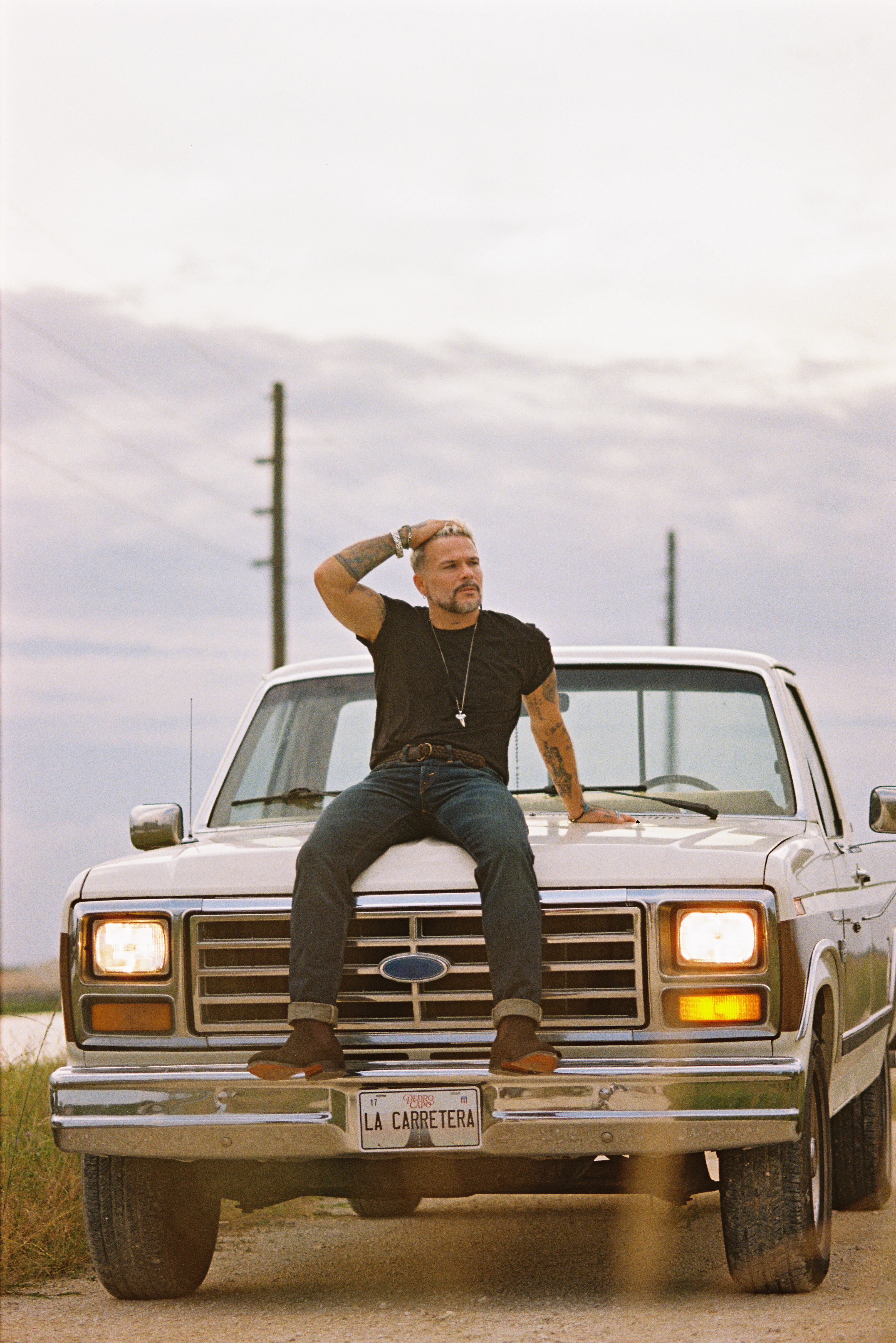 Imágenes promocionales del nuevo disco de Pedro Capó. Se ve al artista subido en la tapa de un carro y al final de un camino sosteniendo su guitarra.