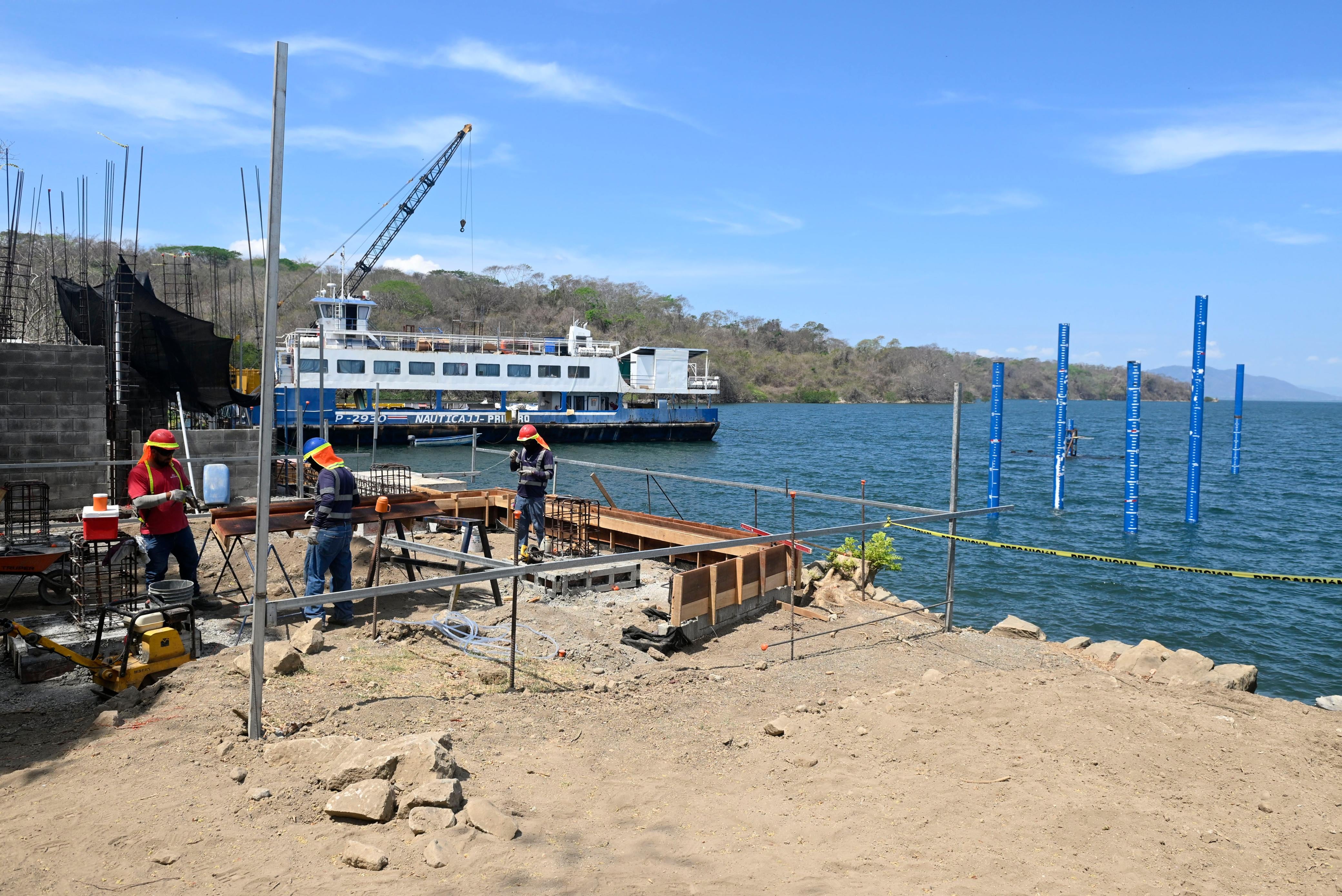 Obreros construyen muelle turístico en isla San Lucas con ferry cercano en Puntarenas y pilotes visibles en el mar.