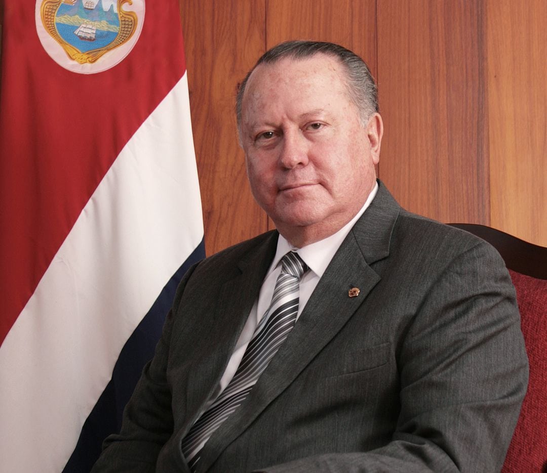 Fotografía del Dr. Luis Paulino Mora Mora, expresidente de la Corte Suprema de Justicia, sentado junto a una bandera de Costa Rica.