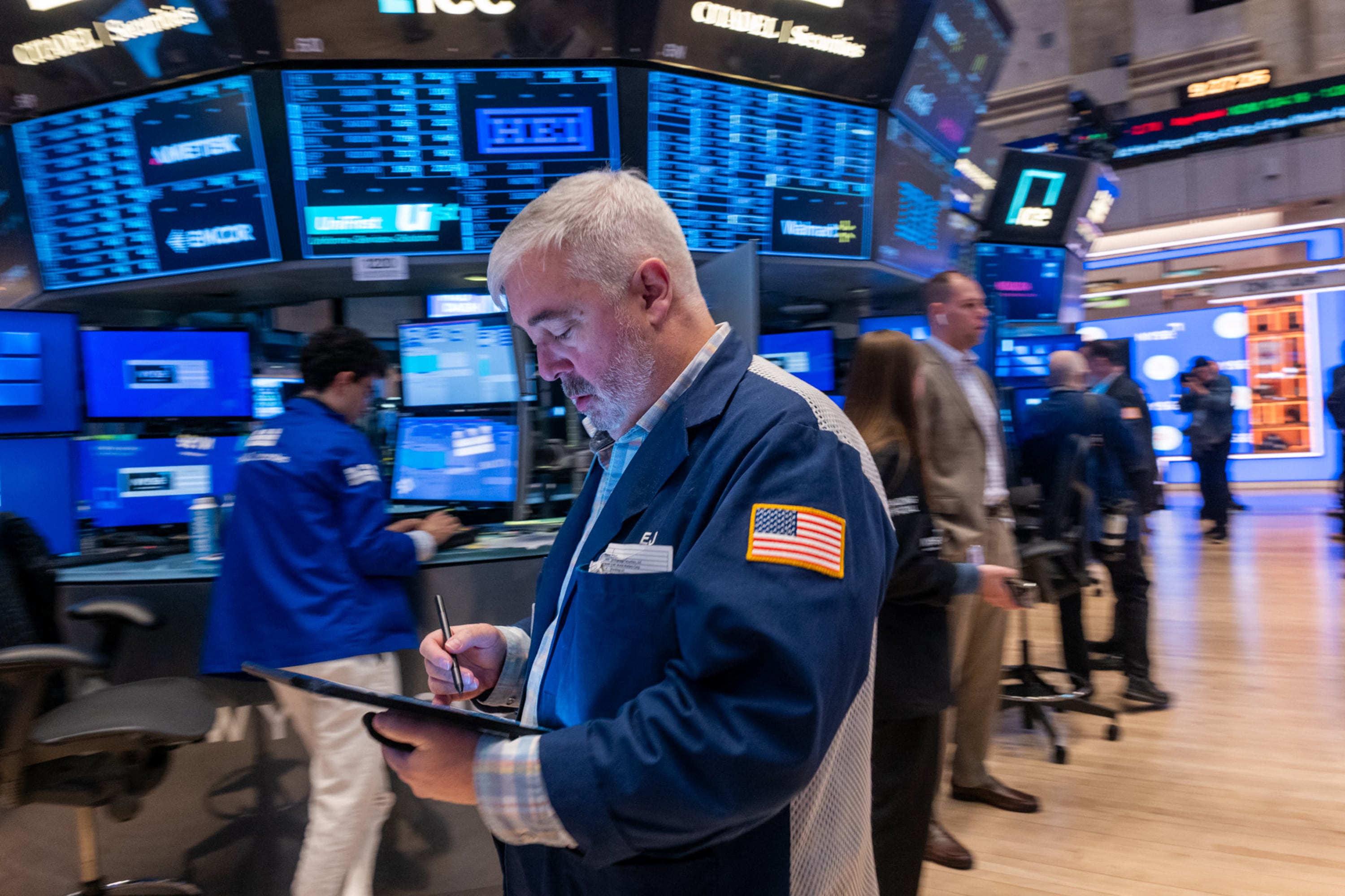 Corredor de bolsa con tablet trabaja frente a múltiples pantallas en la Bolsa de Nueva York, en un contexto de repunte bursátil tras acuerdo comercial entre EE. UU. y China.