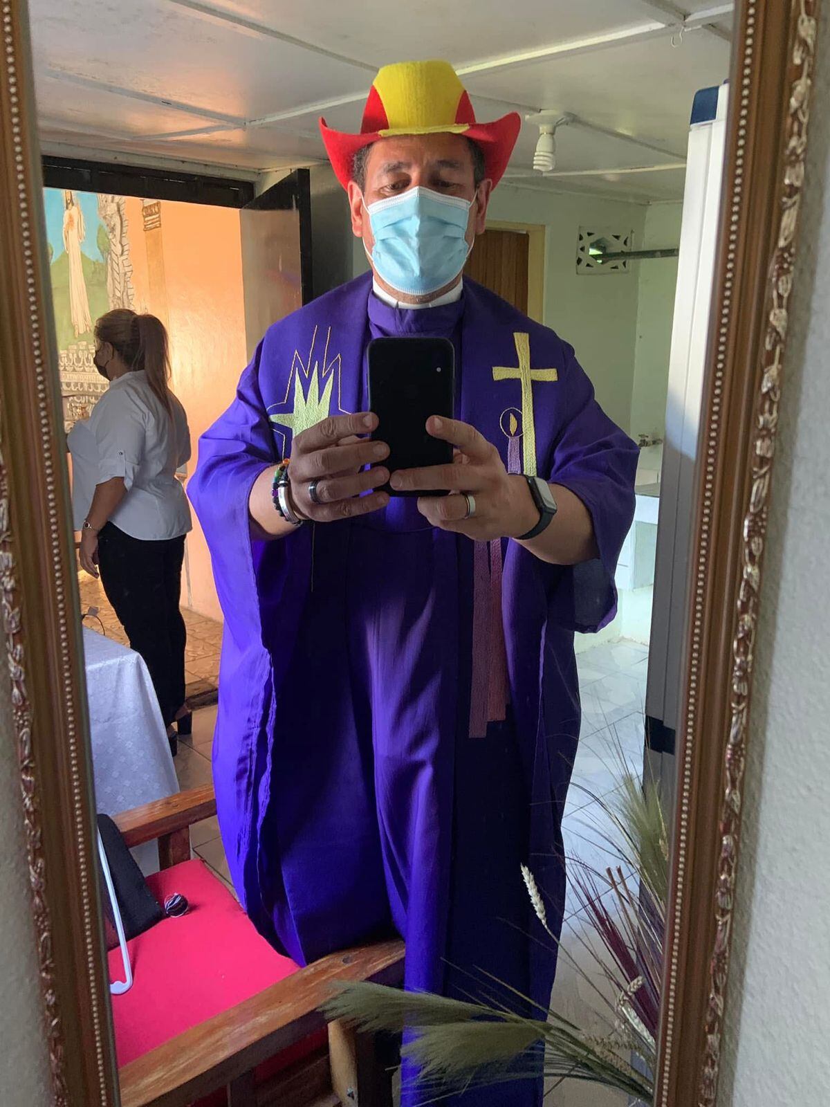 Sacerdote vestido de morado, con sus prendas religiosas, se toma un selfie frente a un espejo. Lleva puesto un sombrero rojo y amarillo.