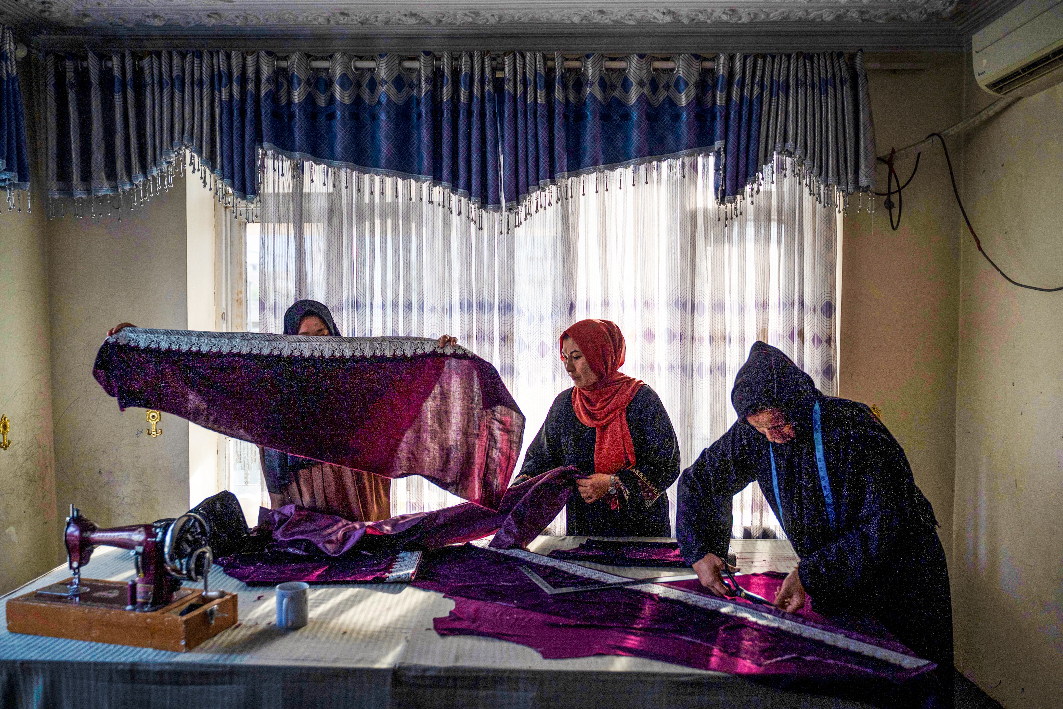Tres mujeres afganas trabajando en un taller de costura, confeccionando prendas de color púrpura bajo la luz natural que entra por una ventana con cortinas.