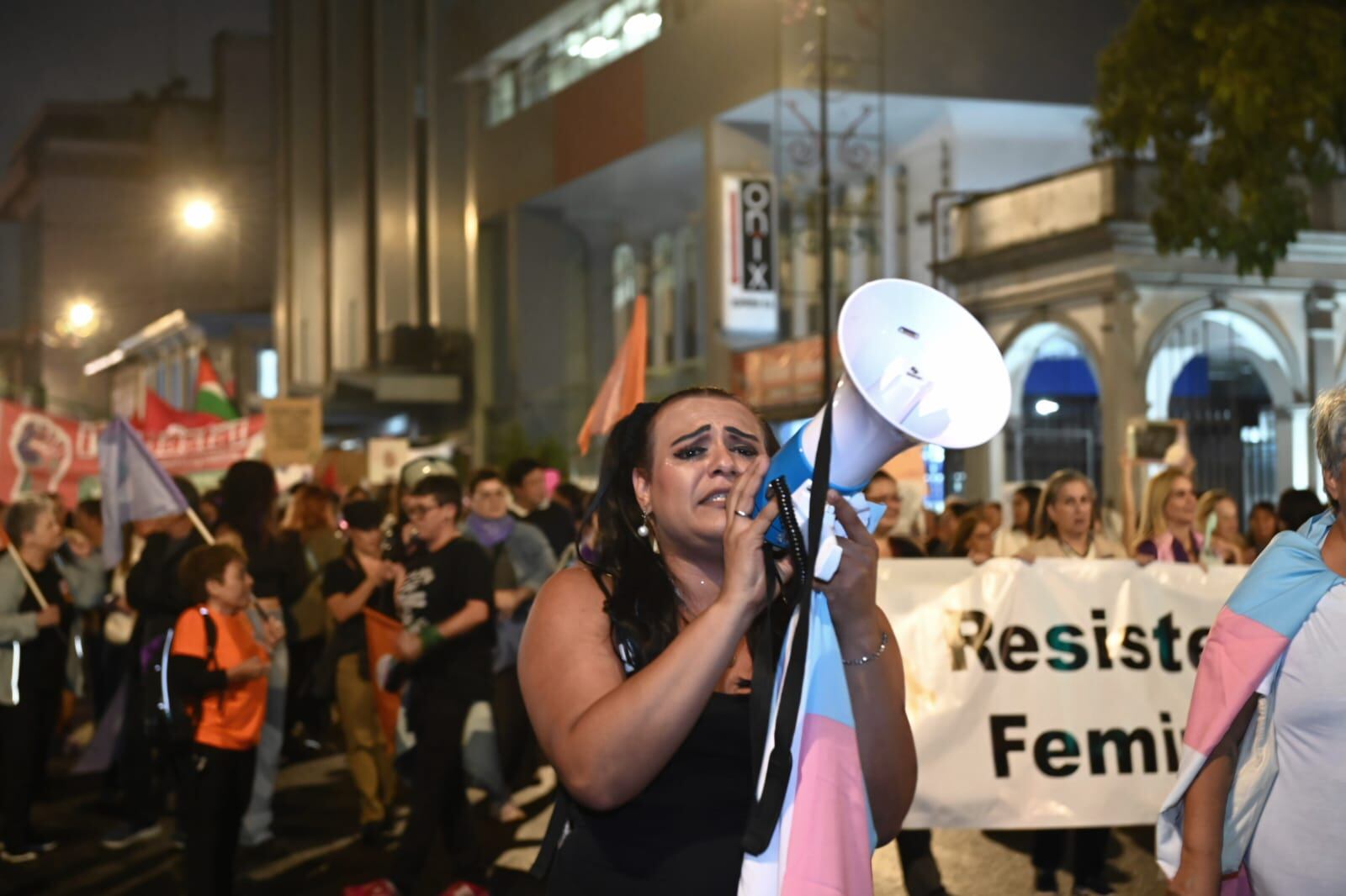Mujer gritando consignas con un megáfono en una marcha nocturna en San José, rodeada de otras personas que sostienen banderas y pancartas contra la violencia de género.